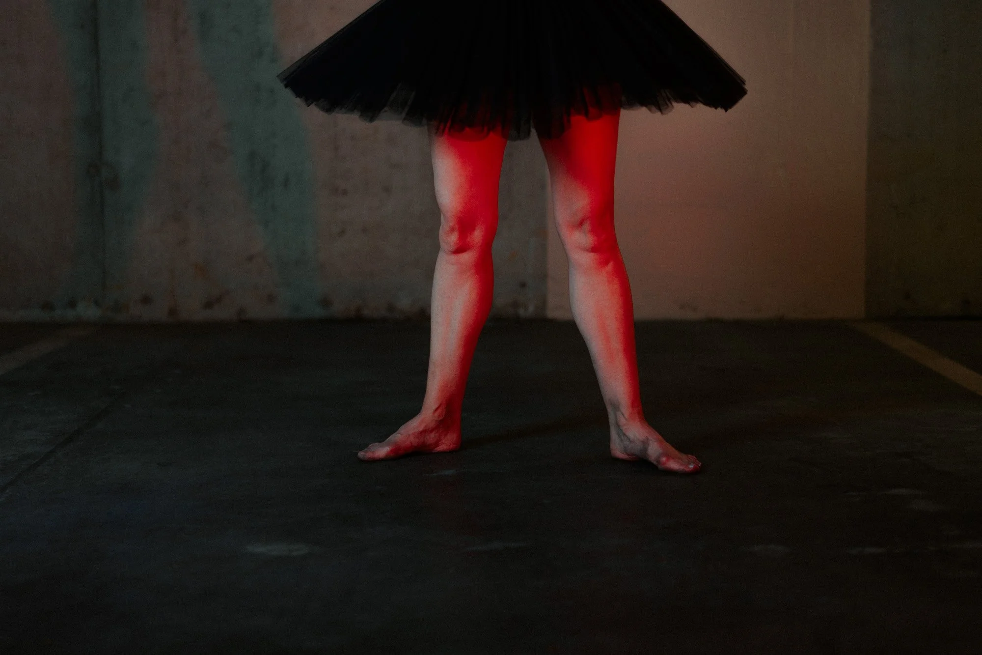 Close-up of a person wearing a black tutu and fishnet stockings, standing barefoot in a dimly lit environment with a textured background.