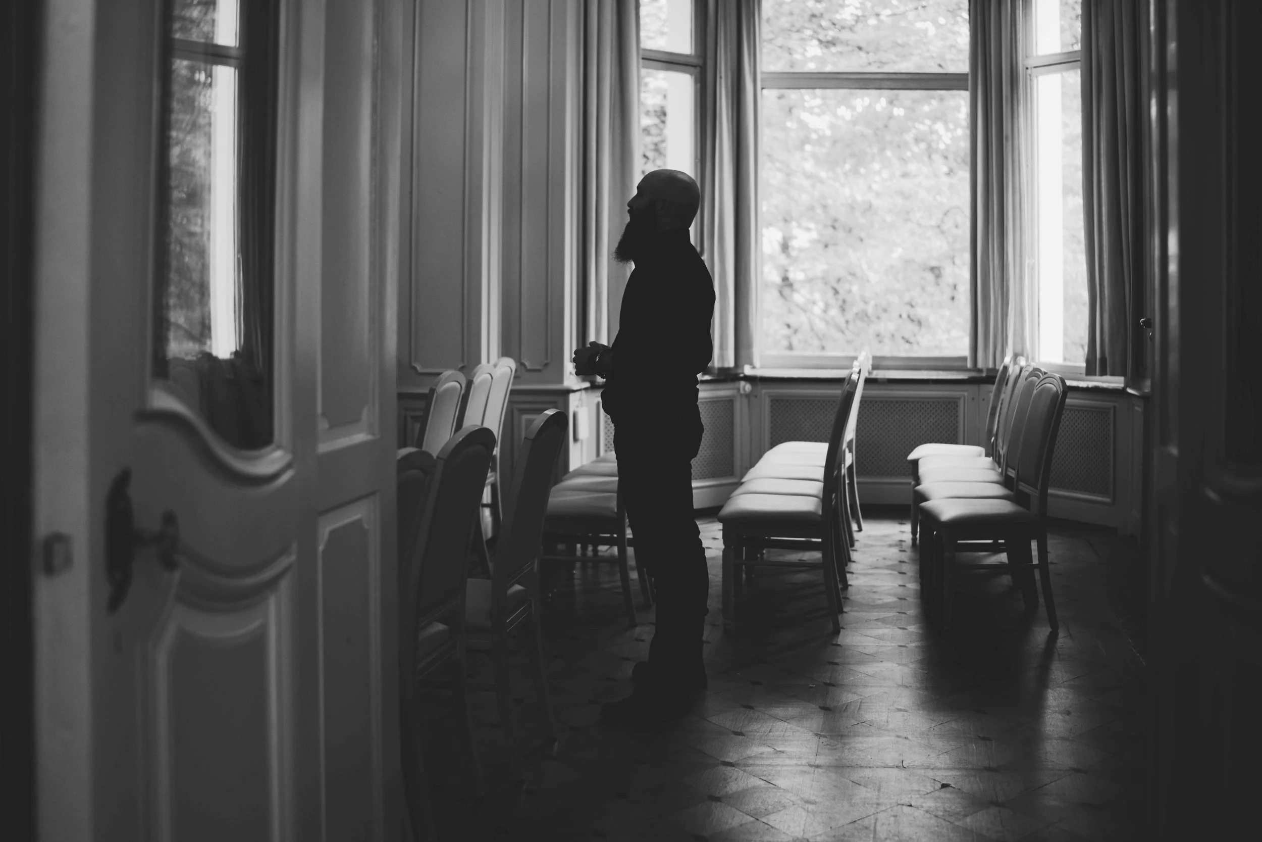 Silhouette of a groom with a beard standing alone in a town hall room with empty chairs, large windows, and draped curtains, in black and white.