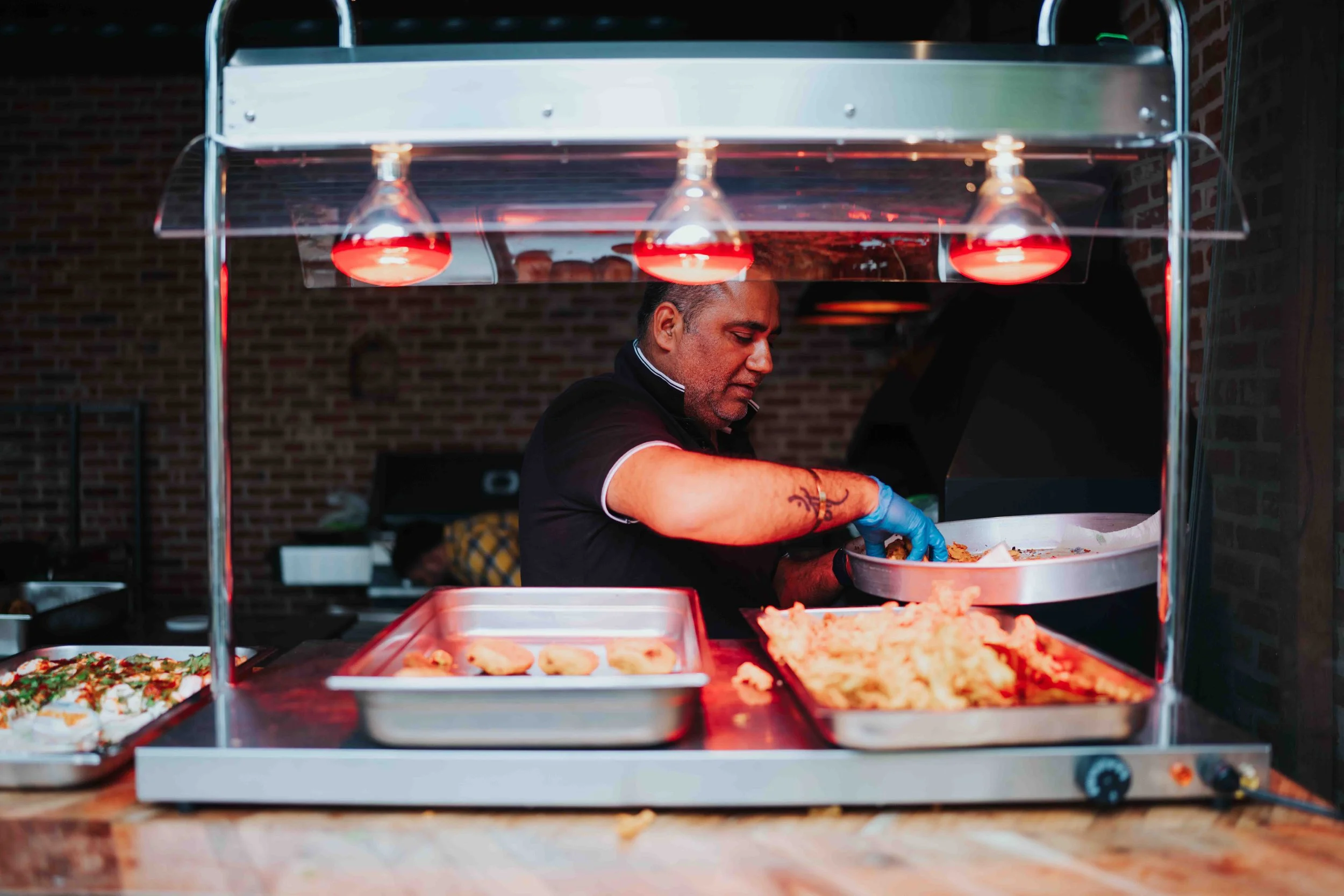 A man wearing a black polo shirt with white trim and blue gloves preparing food behind a food counter with trays of food, in front of a brick wall.