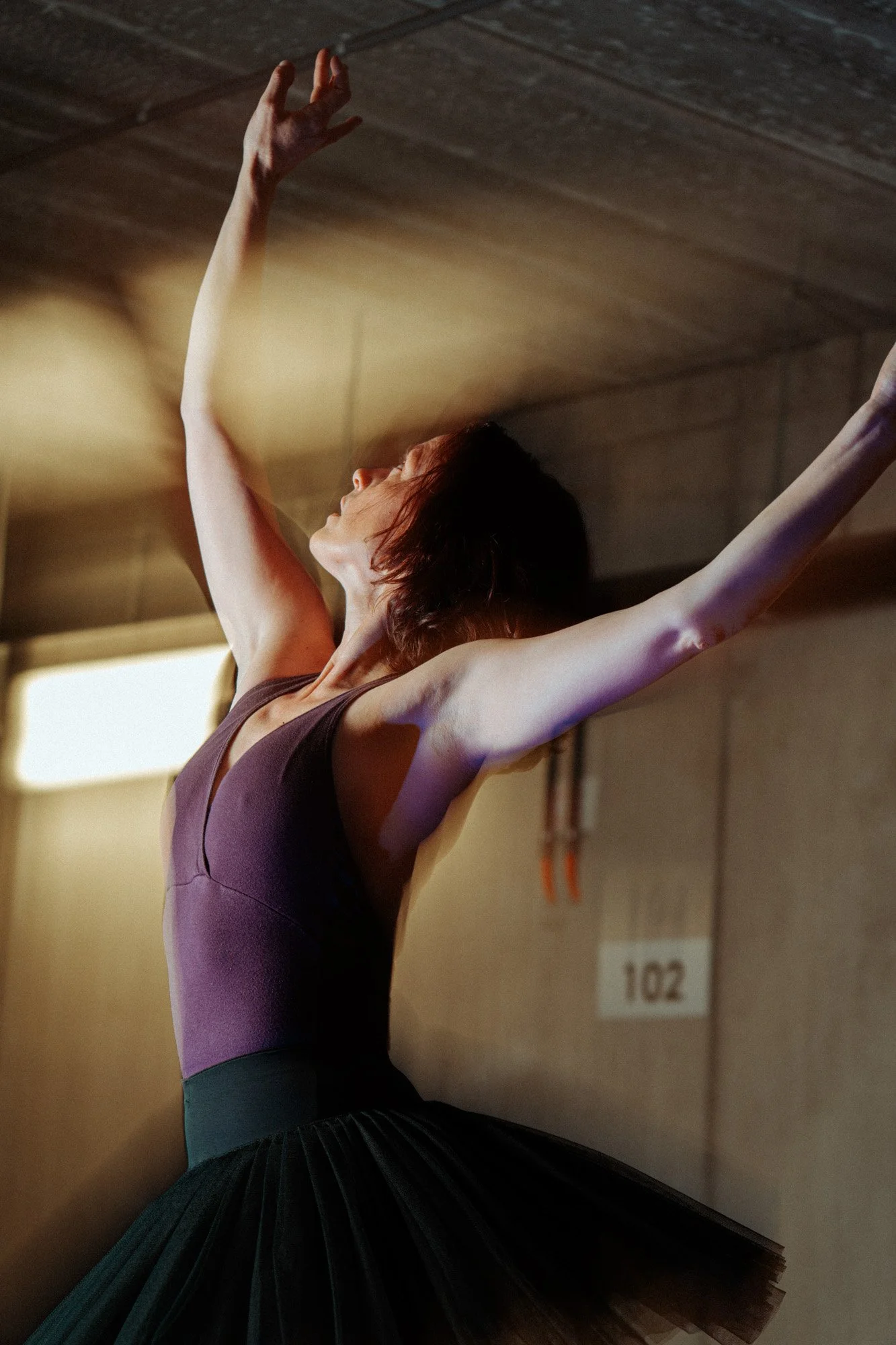 A ballet dancer in a purple leotard and black tutu performing an expressive dance pose in a dimly lit studio.