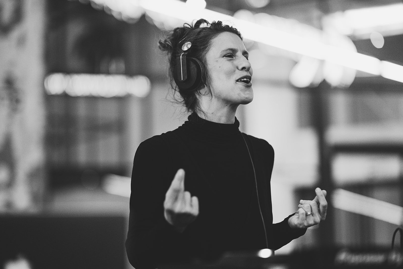 A black-and-white photo of a woman wearing headphones, singing or speaking with her eyes closed and hands raised, in a possibly performance or recording setting.