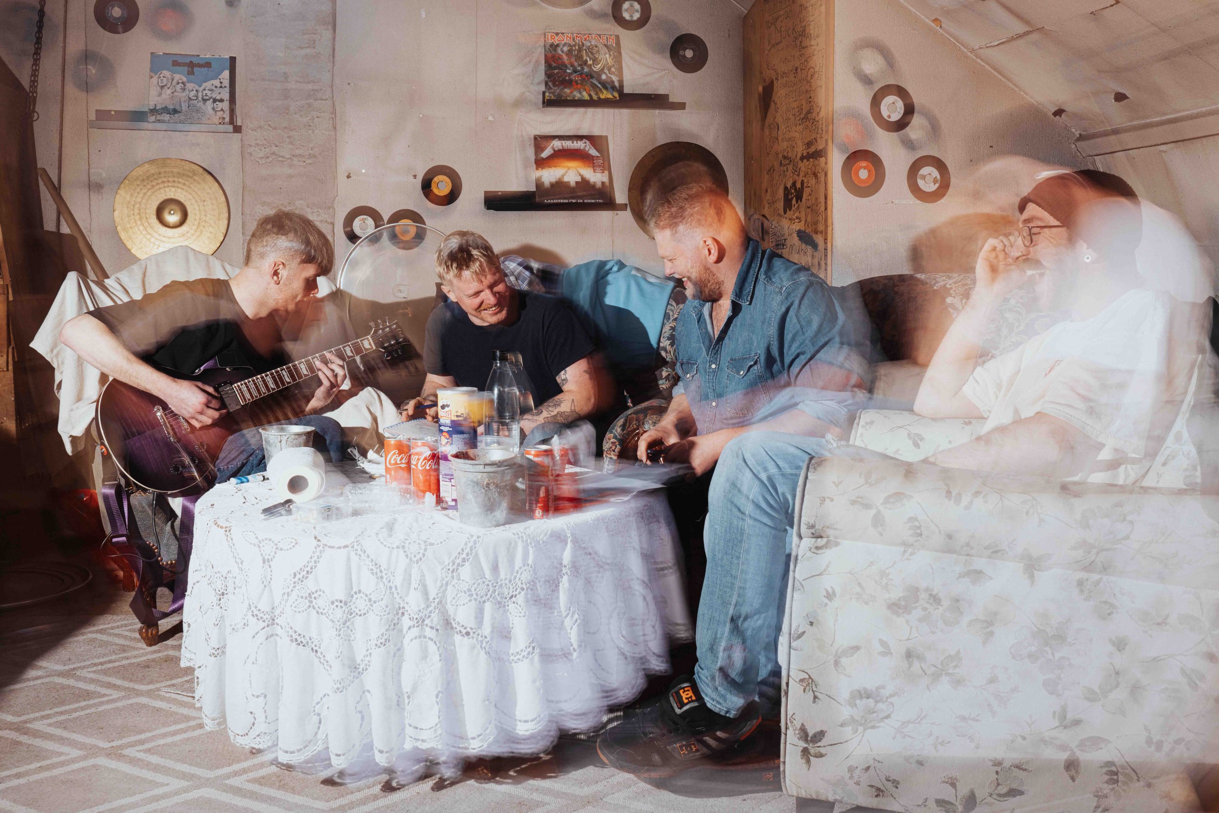 Group picture of the trash-metal band Dread. Four young men sitting around a table in a cozy room, playing guitar, drinking soda, and laughing together, with one of them sitting on a sofa nearby.