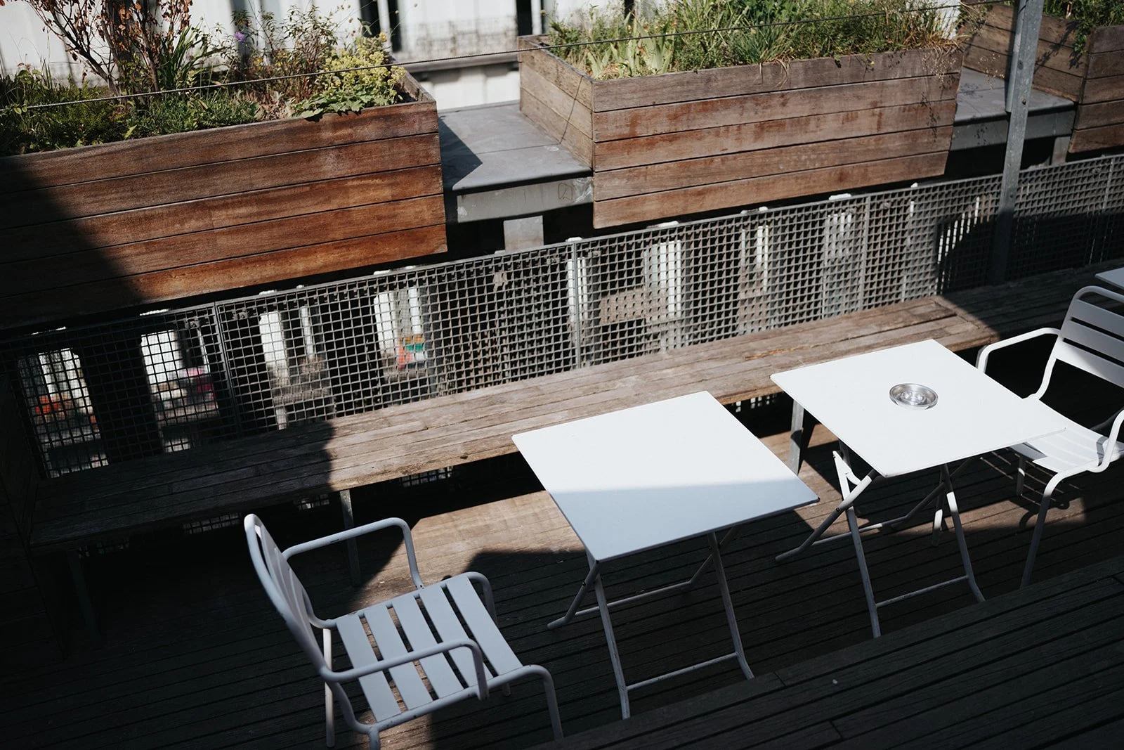 Outdoor patio with wooden and metal fencing, two white square tables, and white chairs, with some shadow and sunlight.