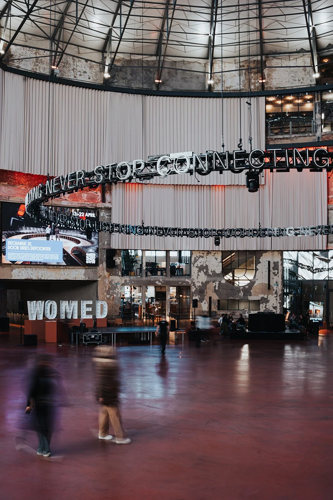 Large indoor space with modern industrial design, featuring a prominent neon sign that reads 'Connecting Never Stop Connecting' hanging from the ceiling. There are people walking around and sitting at tables, with a large 'WOMED' sign on the left sid