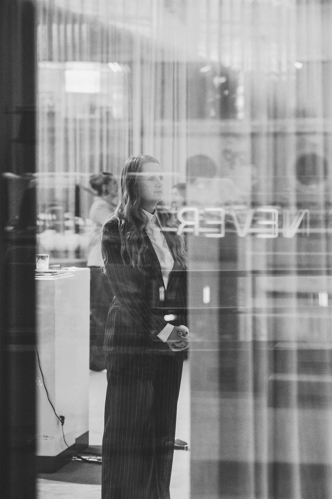 Woman in business suit standing inside a building, looking out through a glass window with the word 'NEVER' reflected on it.