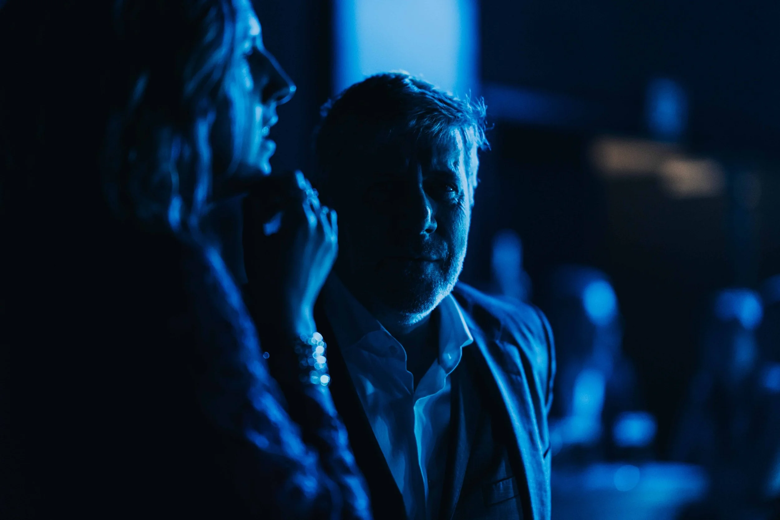 A man and woman in a dark room illuminated by blue light, engaging in conversation, with the woman in the foreground and the man looking towards her.