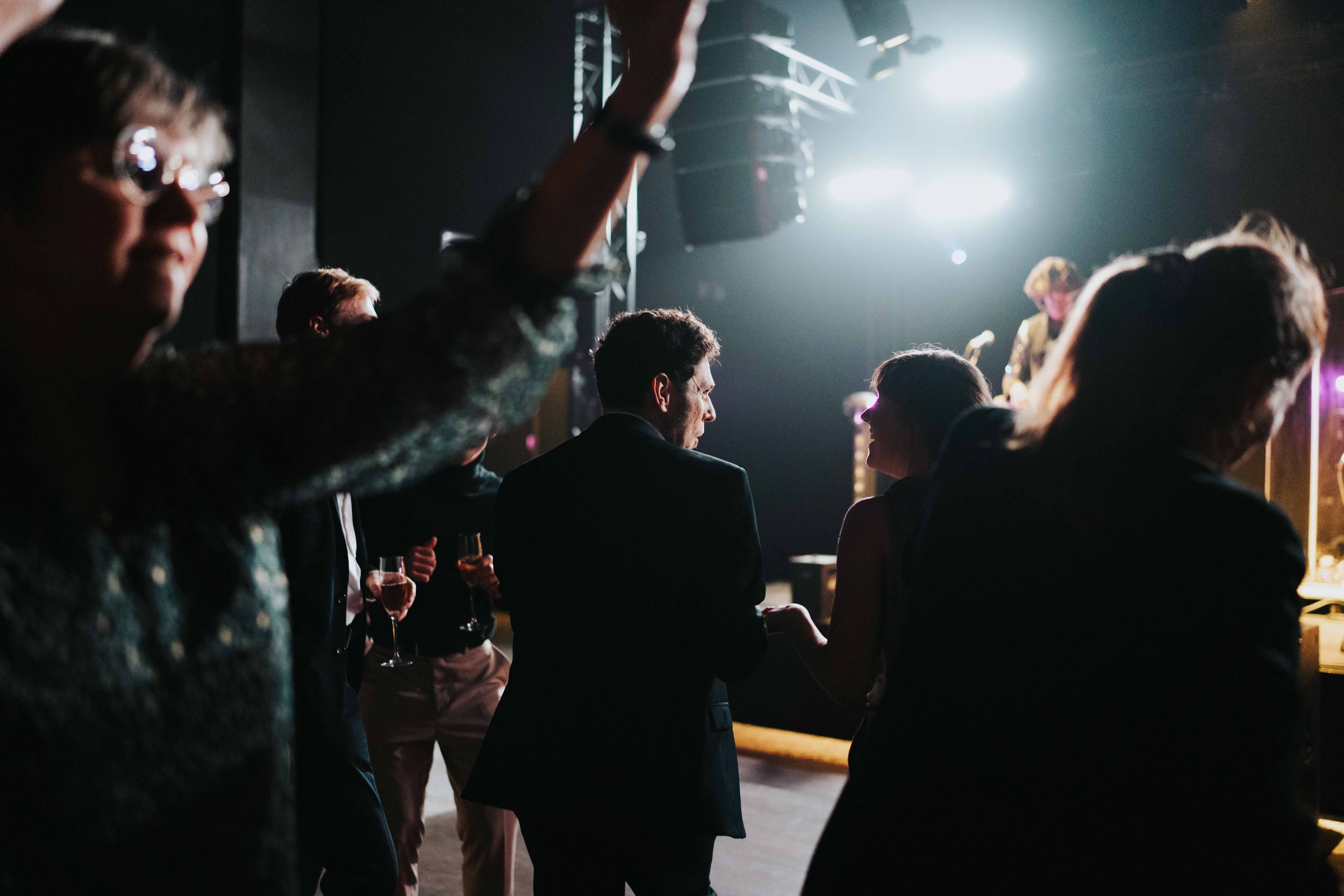 People dancing and socializing at a dimly lit indoor event, with some holding glasses of champagne and others engaging in conversation.