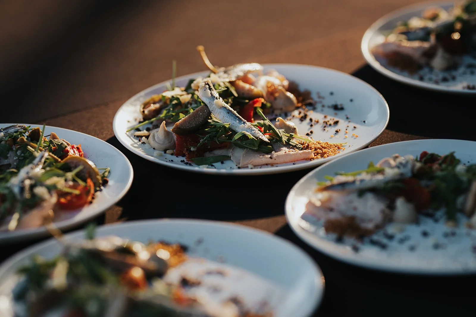 Several plates of salad with greens, vegetables, cheese, and toppings on a dark surface, illuminated by warm lighting.