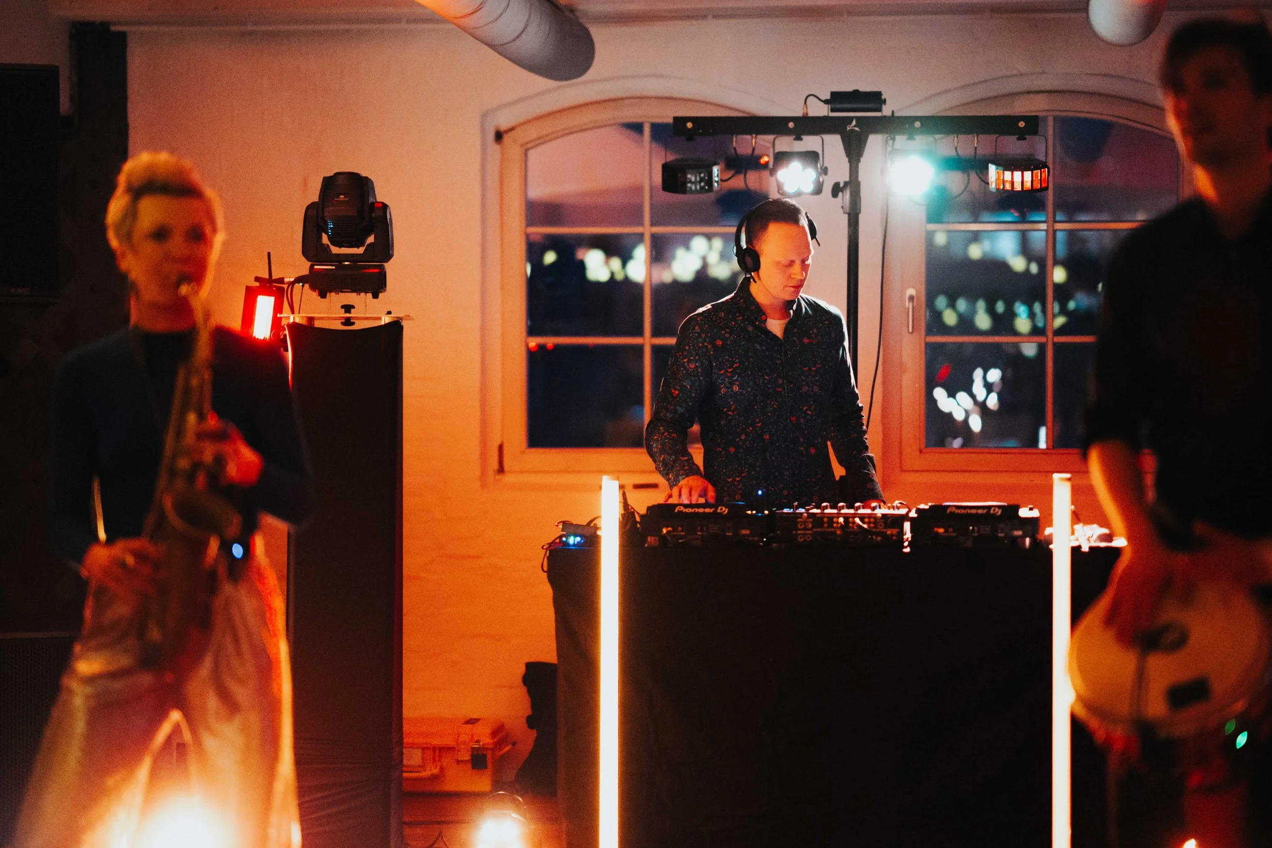 A DJ is performing at a party with a neon-lit dance floor, with two women in the foreground, one holding a saxophone and the other a guitar. The DJ is standing behind a table with equipment, wearing headphones, with a window and city lights in the ba