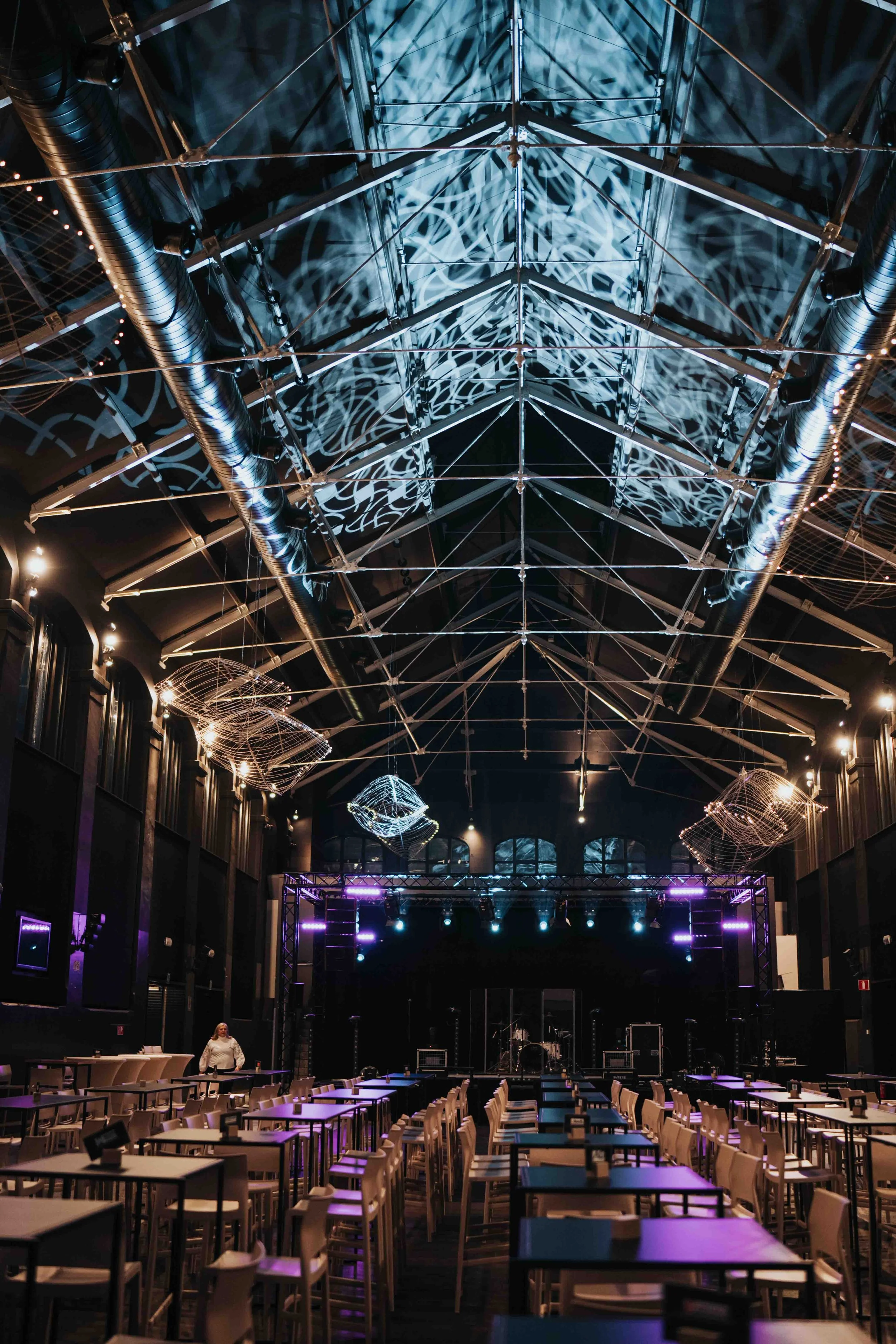 An indoor concert venue (Oude Vismijn Ghent) with a stage, seating, and intricate metal decor hanging from the ceiling, illuminated by purple and blue lights.