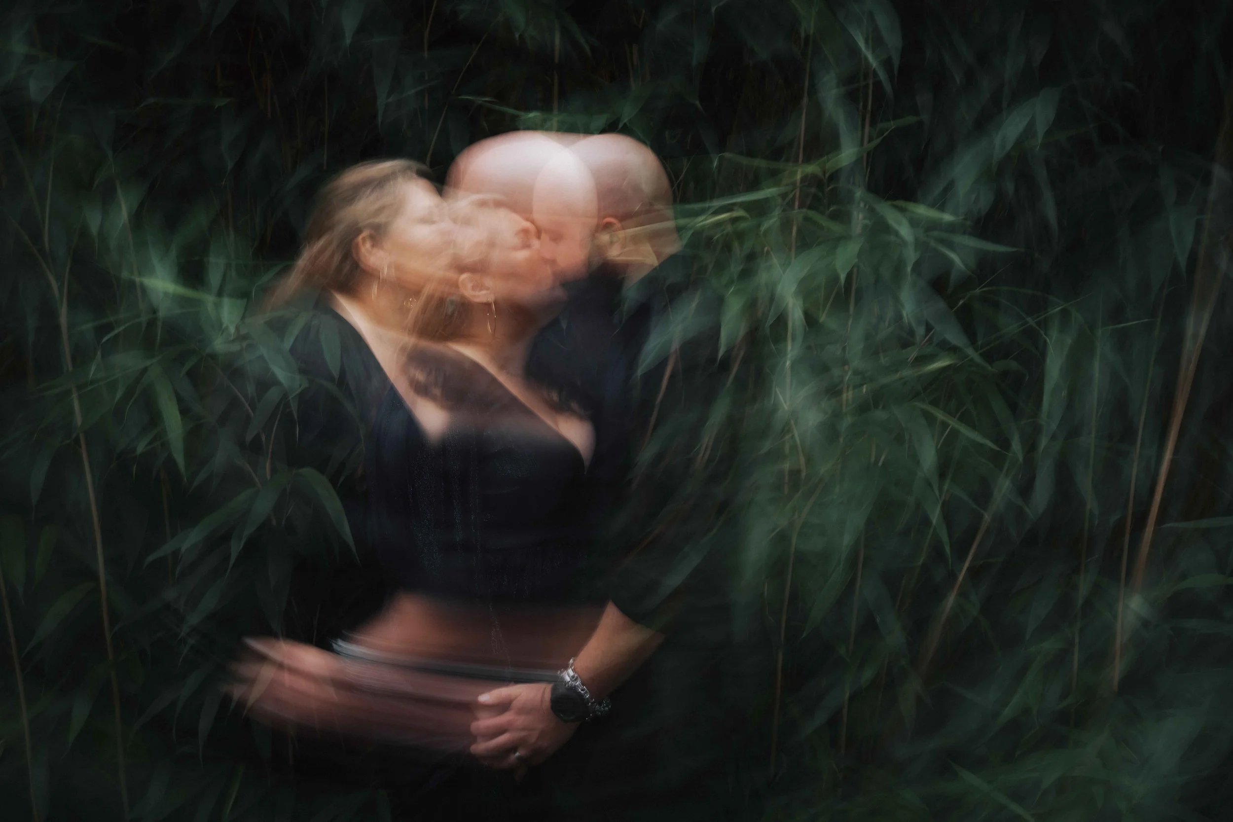 A blurred photo of a couple kissing, surrounded by tall green plants or grass.