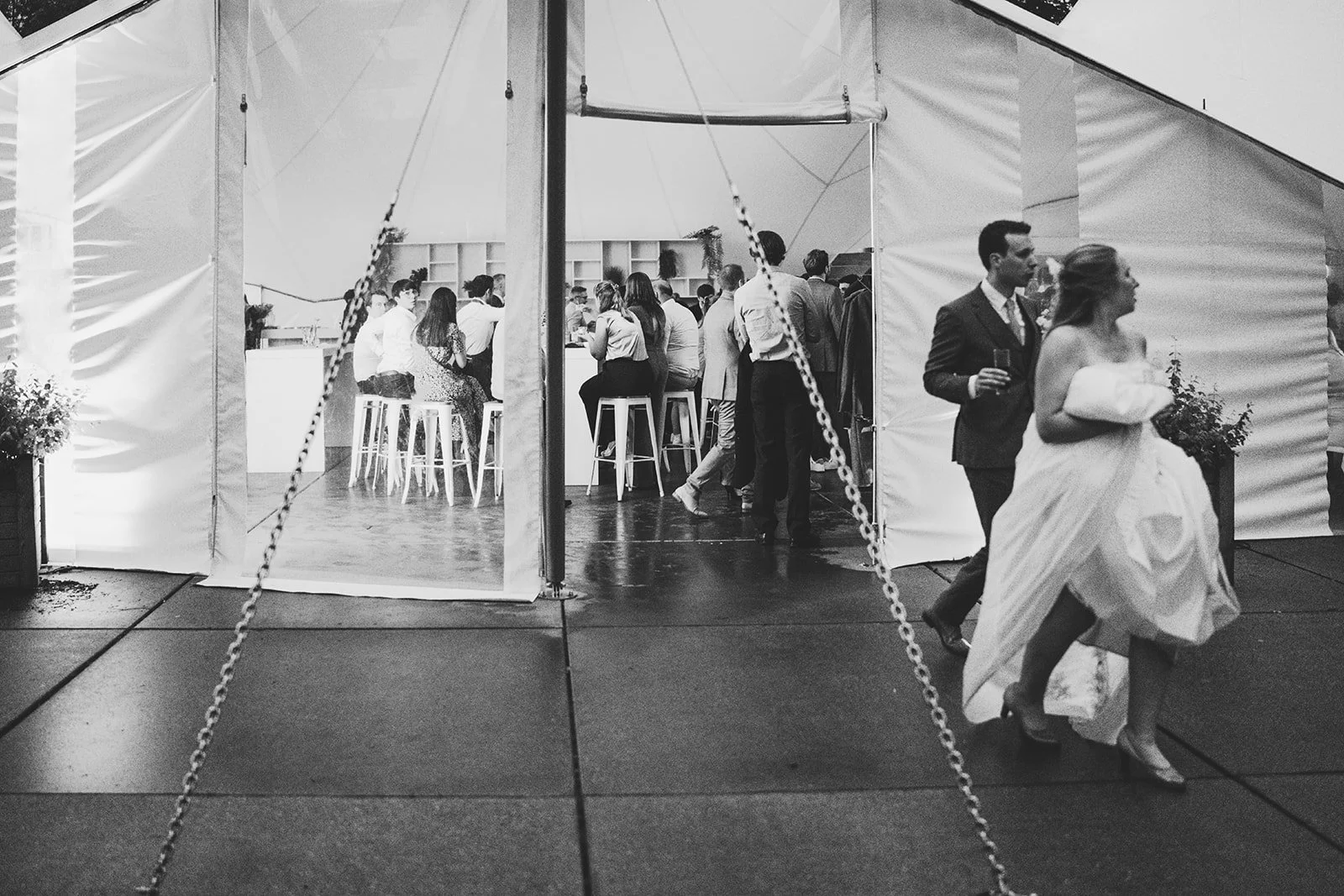 A black and white photo of a wedding reception tent with guests mingling inside and outside, including a bride and groom walking out, with the bride holding a bouquet.
