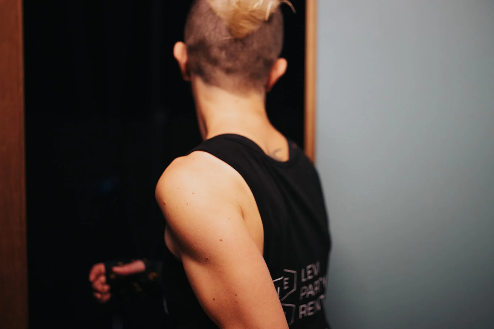 A woman with short, mohawk hairstyle and sleeveless black shirt, walking through a door with their back to the camera, exposing her muscles.