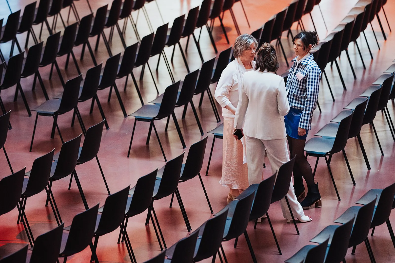 Three women standing and talking in an auditorium with many empty black chairs arranged in rows.