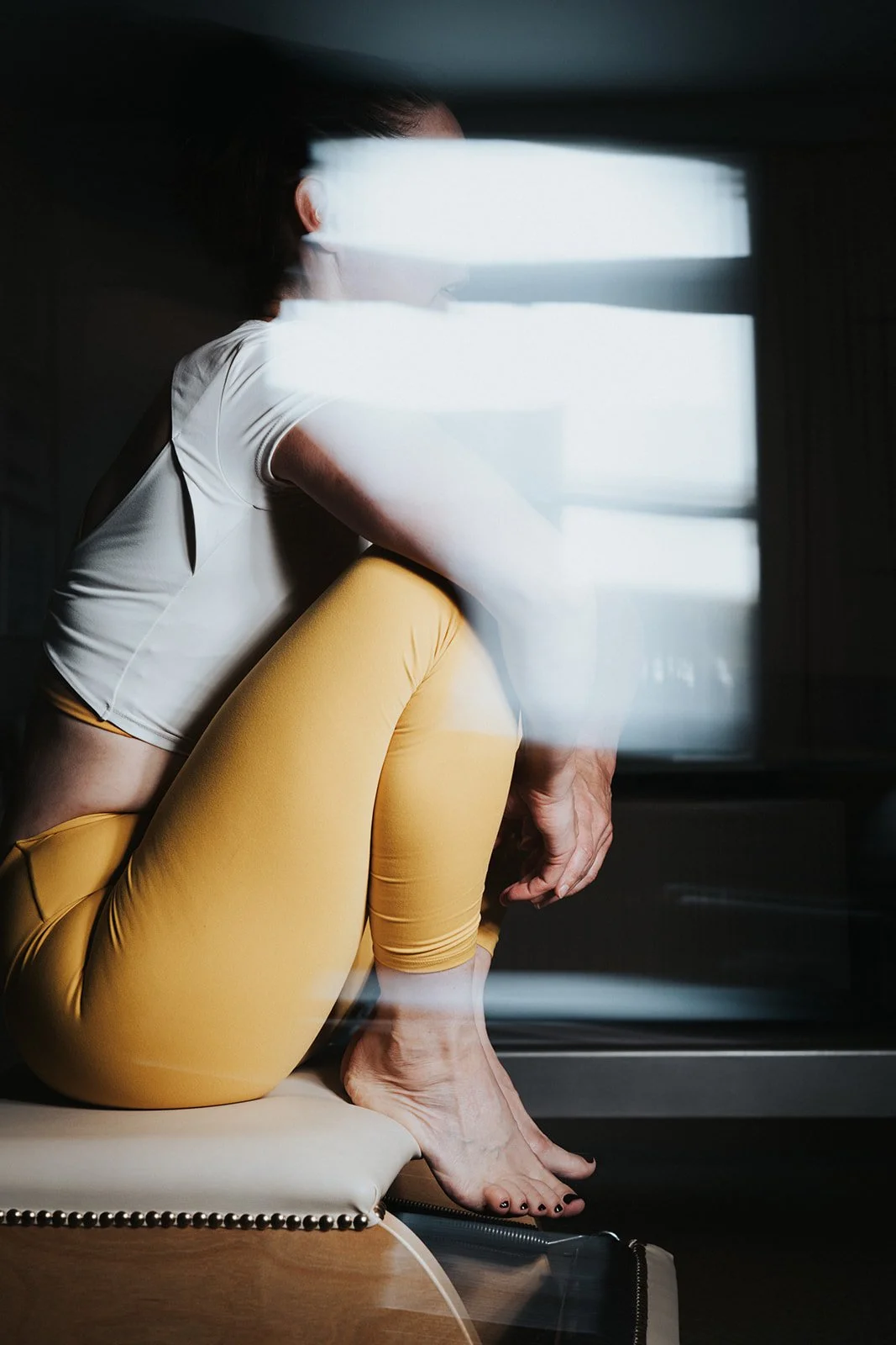 A person with dark hair sitting on a beige chair, wearing a white short-sleeve top and yellow leggings, viewed through a window with horizontal blinds.