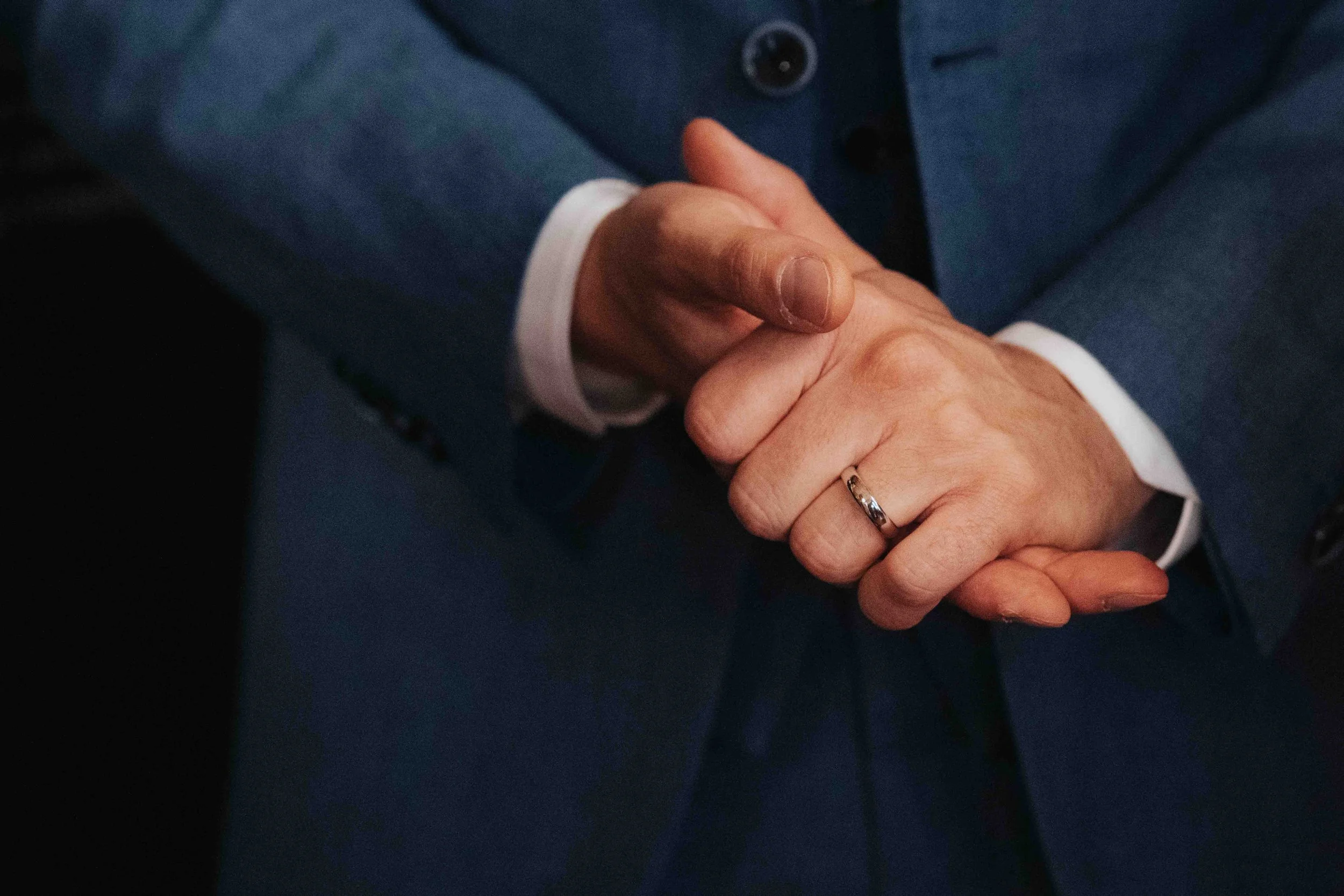 Close-up of a man wearing a blue suit with a white shirt, clasping his hands together, with a wedding ring visible on his ring finger.