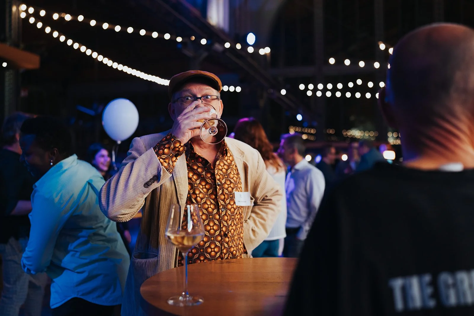 A man with glasses, wearing a patterned shirt and beige blazer, drinks wine at an outdoor social event at night, with string lights overhead and a group of people in the background.