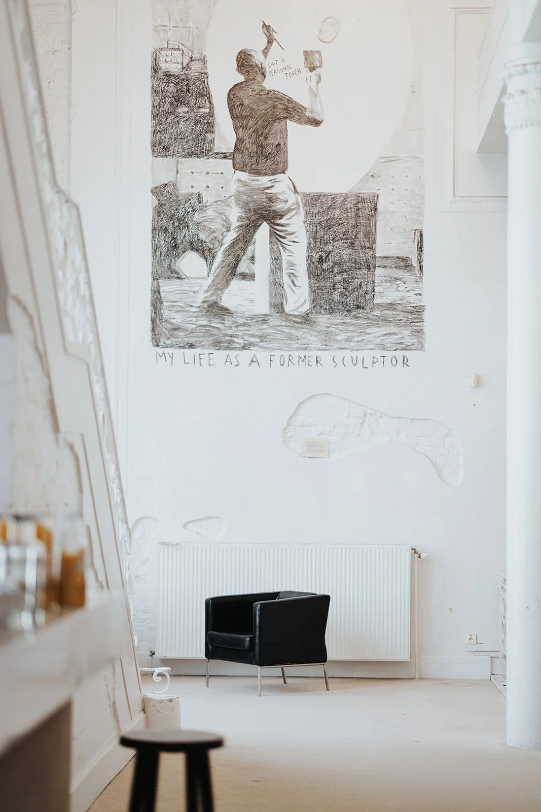 A large wall mural of a man sculpting, with a caption that reads 'My Life as a Former Sculptor,' in a bright, minimalist interior space with a black chair, a white radiator, and some exposed brick patches on the wall.