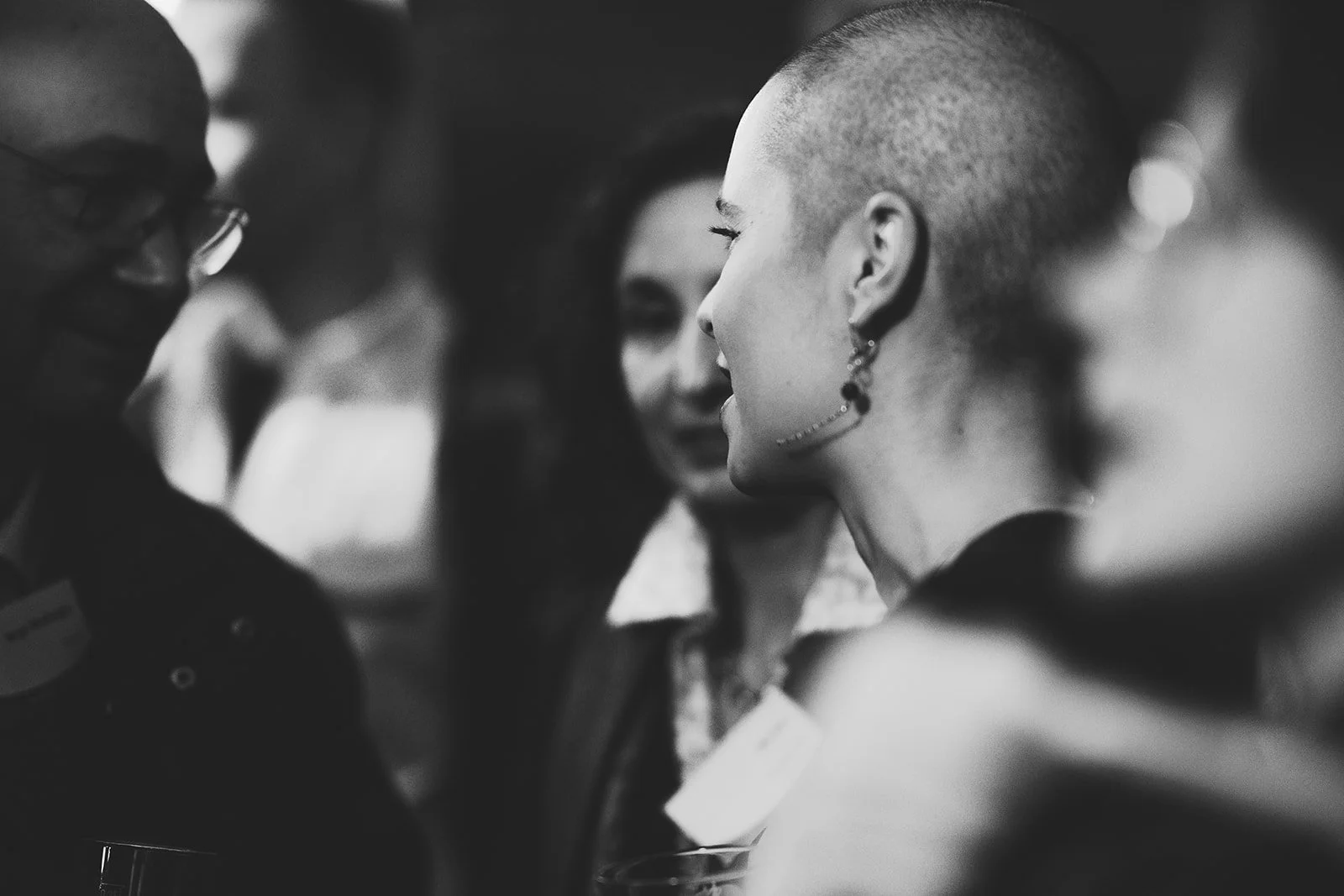 A black and white photo of multiple people talking at a social gathering, with focus on a woman with a shaved head and earrings in the center.