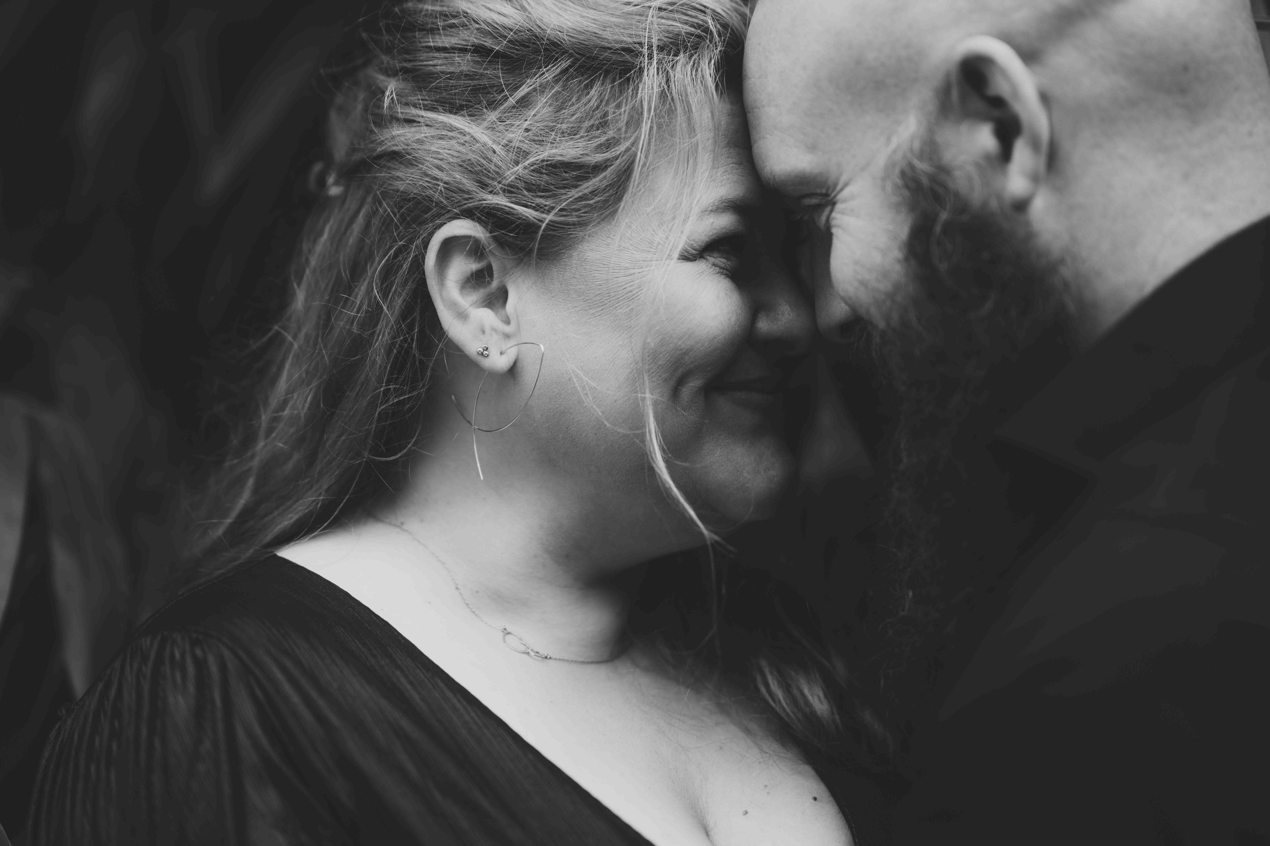 A close-up black and white photo of a man and woman with foreheads touching, smiling with their eyes closed. The woman has earrings and a thin necklace, and the man has a thick beard.