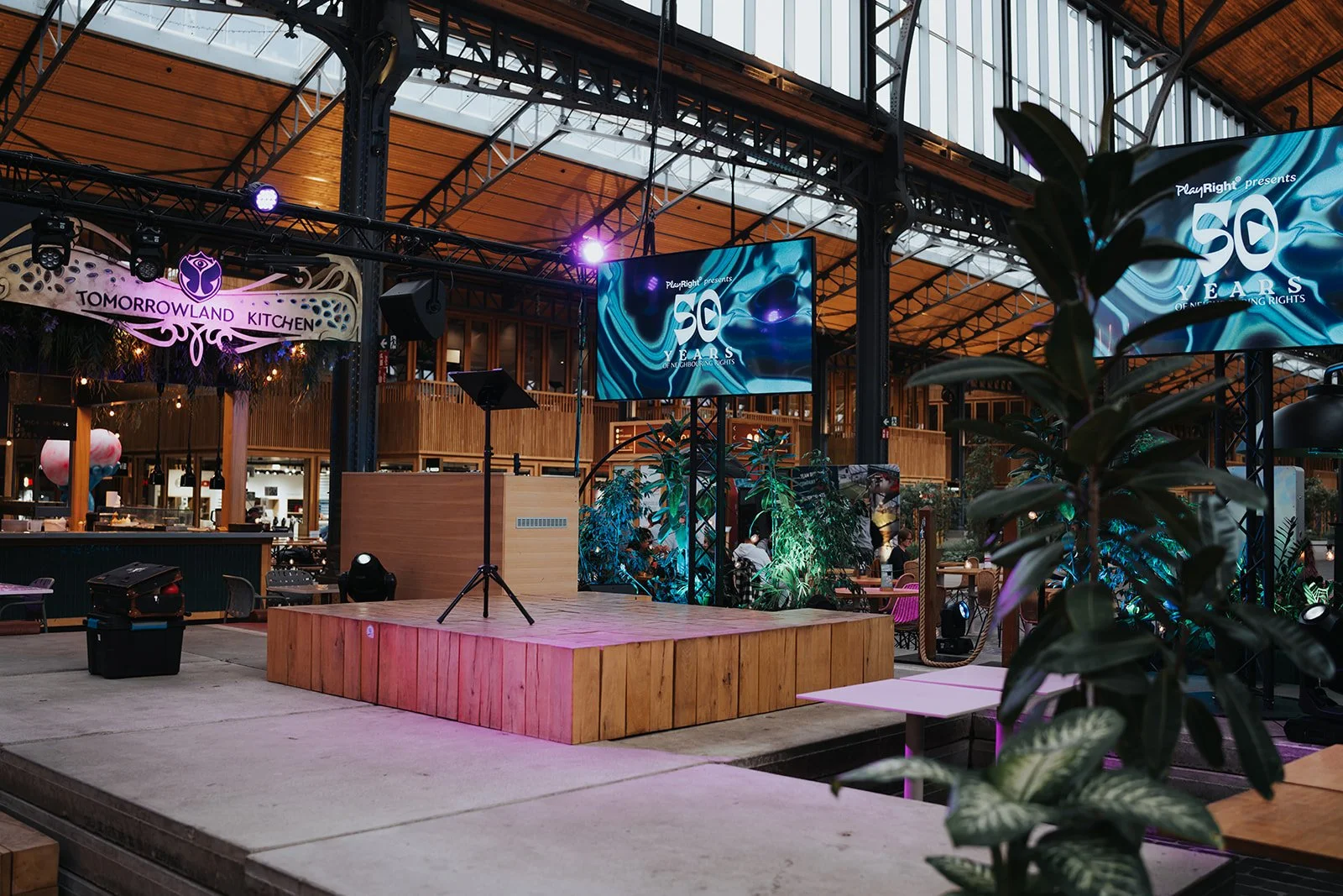 Indoor event space with a small stage featuring a wooden platform, surrounded by large digital screens displaying '50 Years of Human Rights,' with a sign reading 'Tomorrowland Kitchen' in the background, containing plants and seating areas.