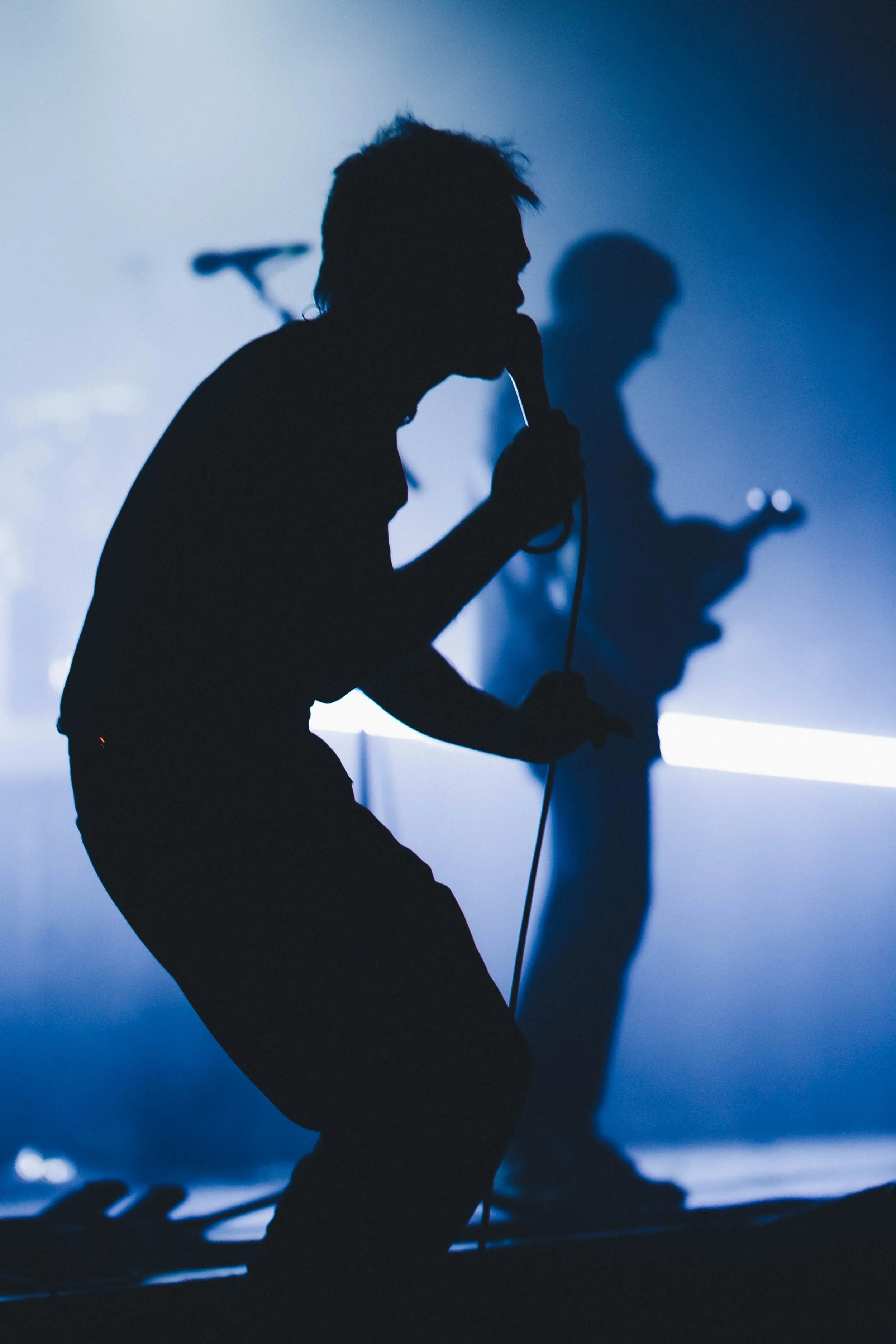 Silhouette of singer Rou Reynolds of Enter Shikari with a microphone on stage, with a guitarist in the background, all against a moody blue lighting backdrop.