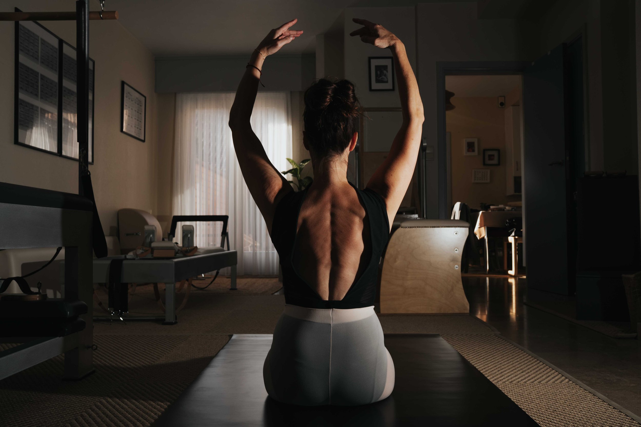 A woman practicing floor ballet indoors, sitting on a mat with arms raised and head turned to the side, in a dimly lit room.