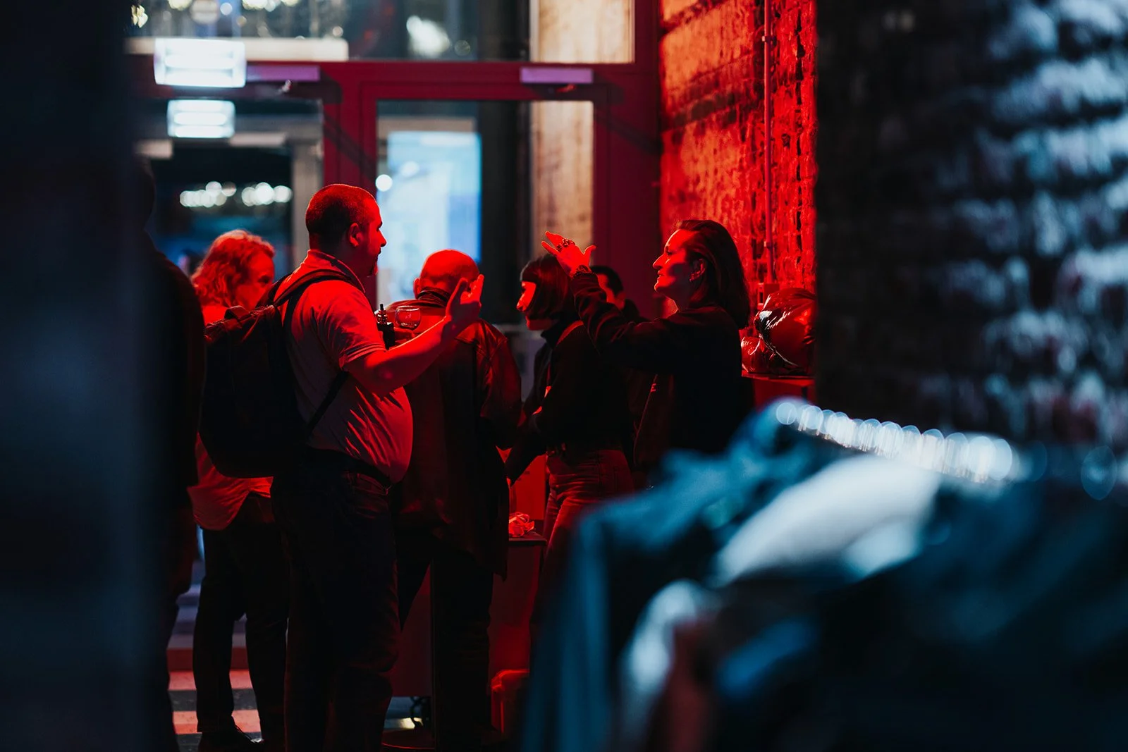 People socializing and engaging in conversations in a dimly lit bar or club with red lighting, brick wall, and large windows in the background.
