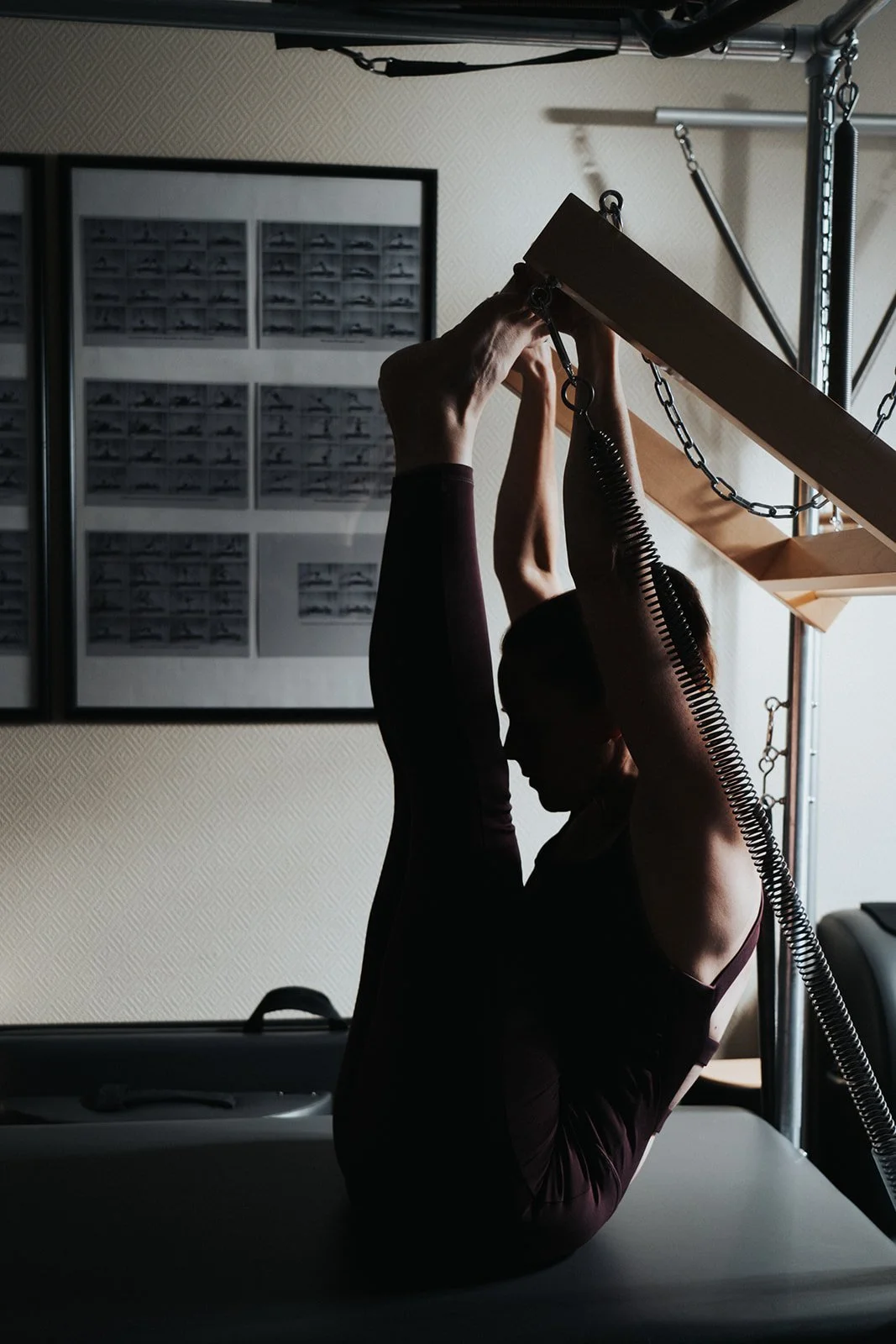 A woman performing a Pilates exercise on a reformer machine, silhouetted against a light source in a gym or fitness studio.