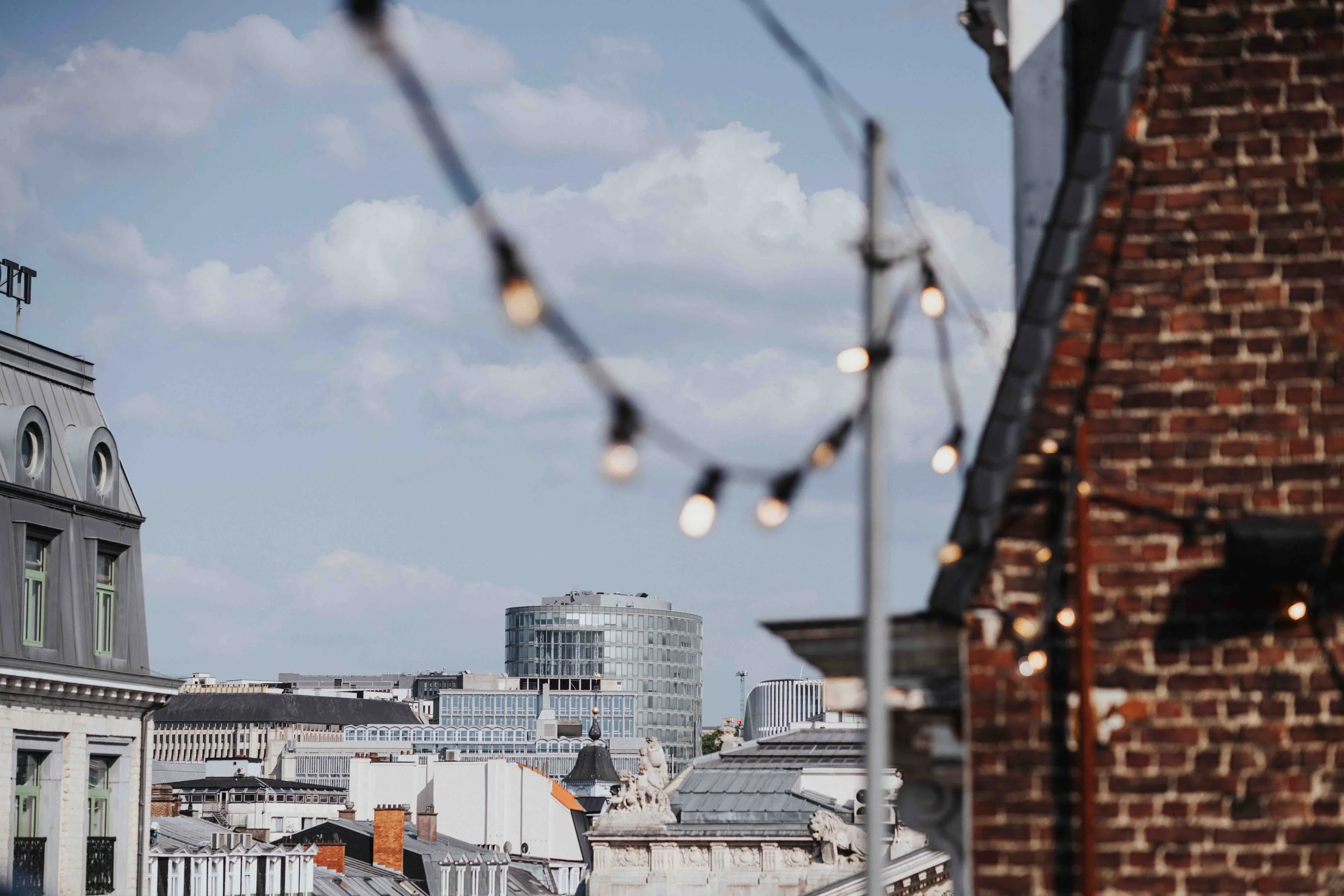 Cityscape view of rooftops with a string of outdoor lights in the foreground, brick building on the right, modern glass building in the background, partly cloudy sky.