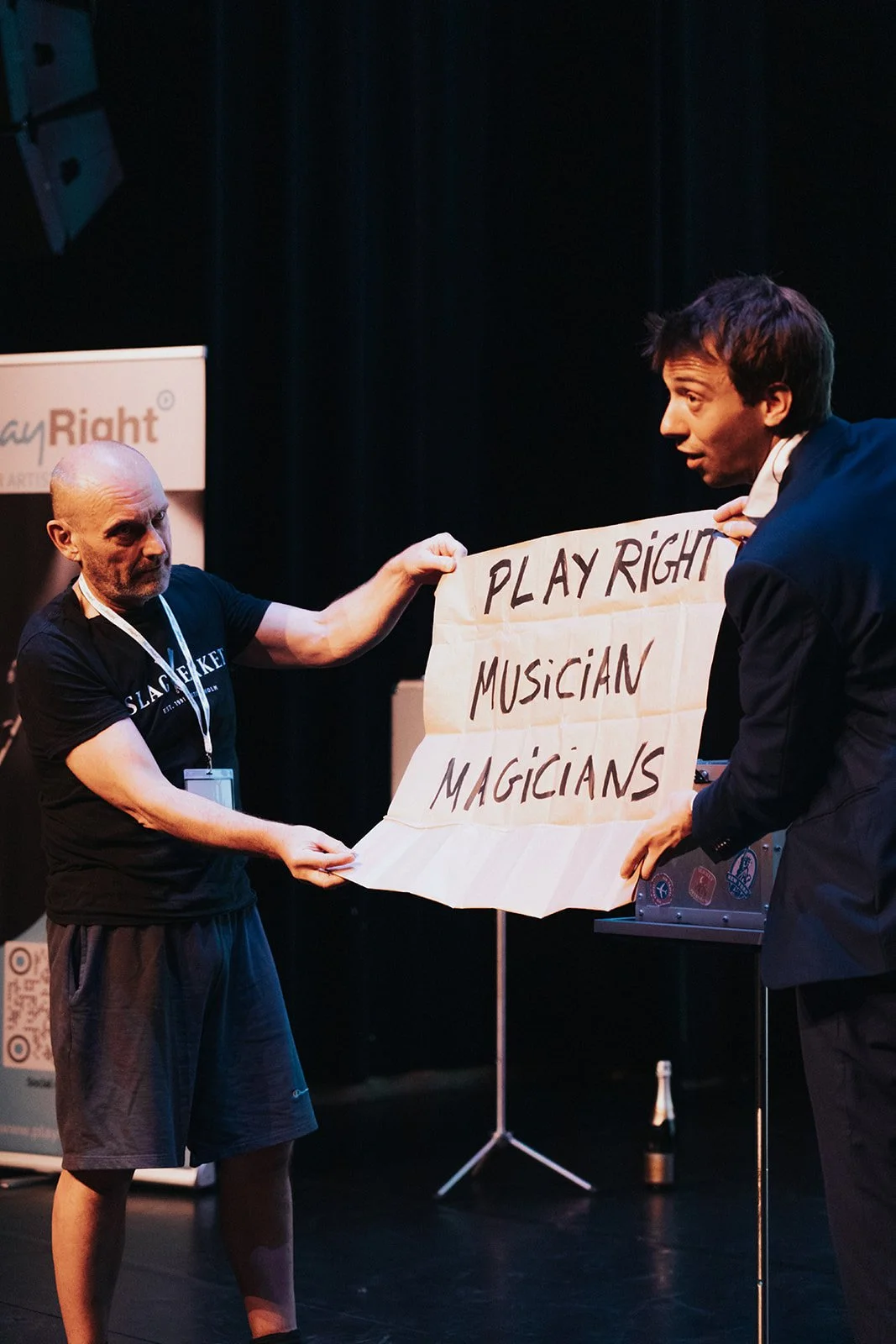 Two men on a stage holding a large paper sign that reads "PLAY RIGHT MUSICIANS MAGICIANS." The man on the left is bald, wearing a black T-shirt and shorts, looking at the other man. The man on the right has dark hair, wearing a blazer, and is looking