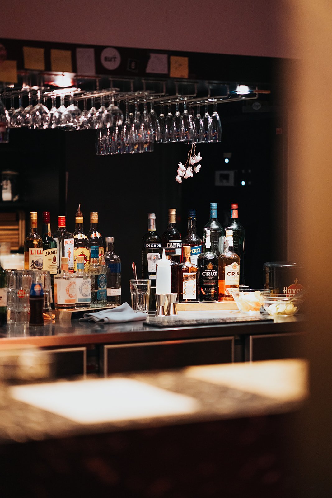 Bar with various liquor bottles, upside-down wine glasses hanging above, and a bowl of food on the counter, with a dark background.