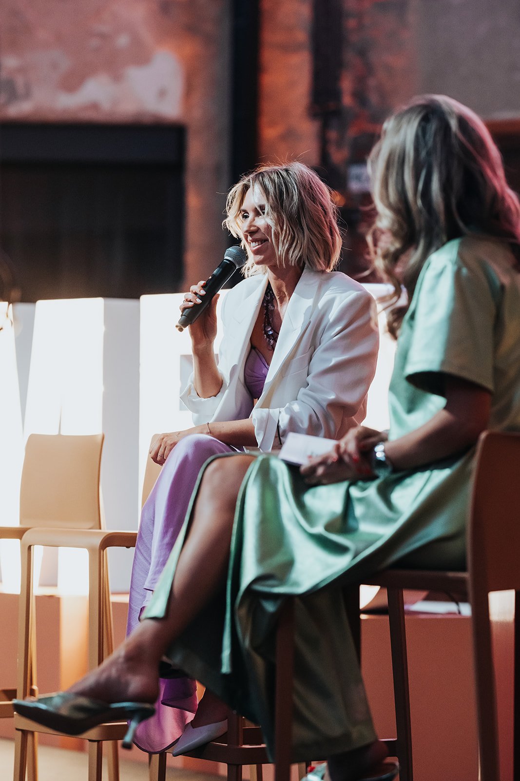 Women participating in a panel discussion, one woman speaking into a microphone and the other woman holding a notepad, seated in a studio or event space with warm lighting and exposed brick wall.