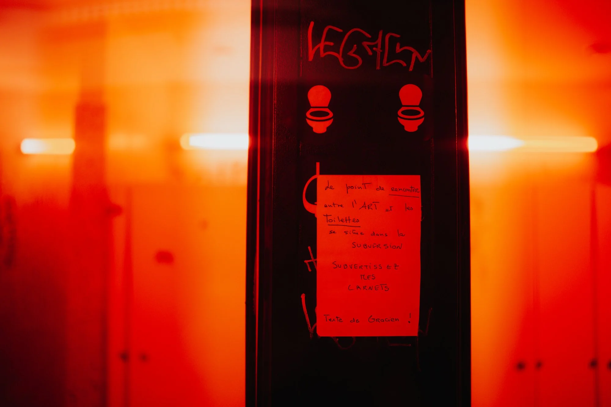 A black vertical sign with red text and symbols, including a note written in French, against a brightly lit orange background.