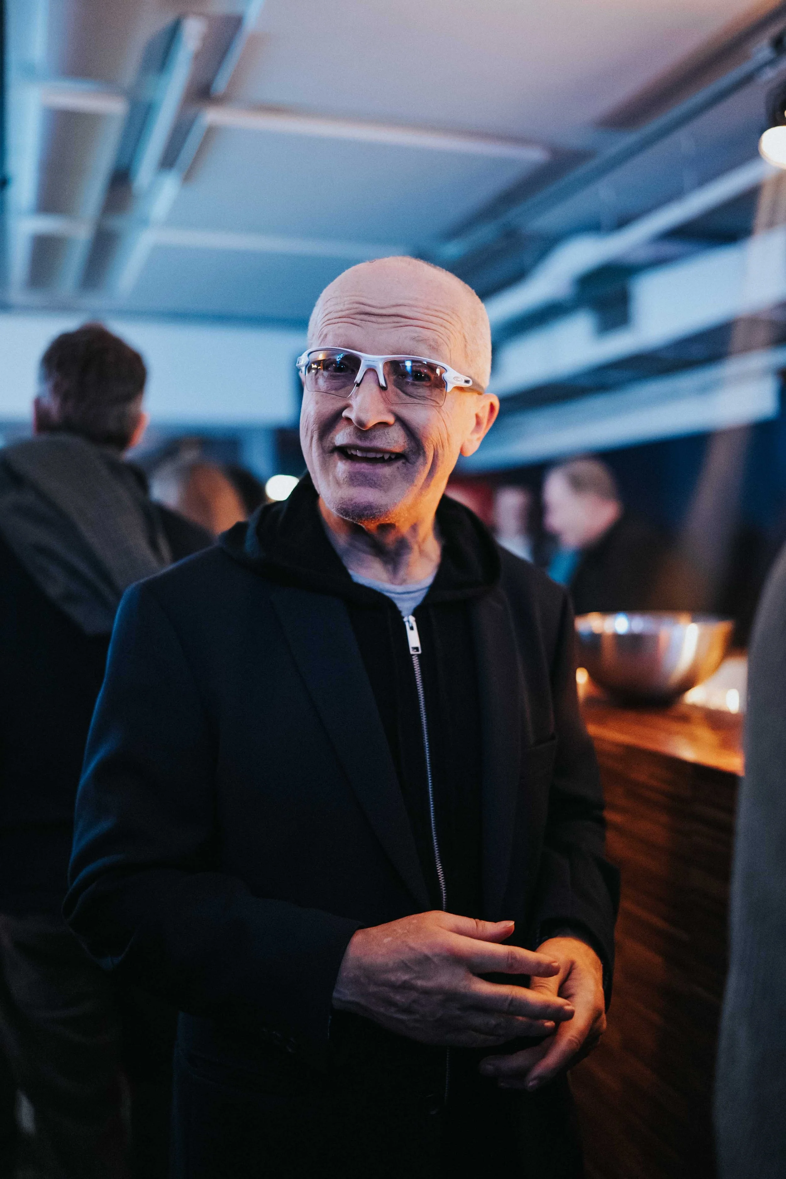 An older man with glasses, wearing a black hoodie and jacket, smiling and talking at a social gathering.