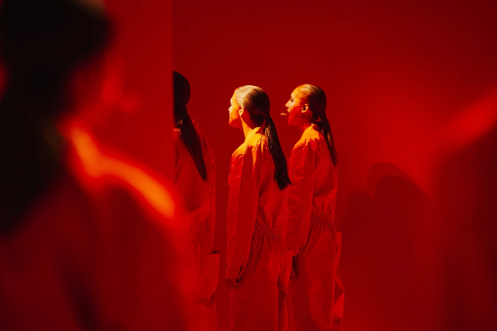 Four women of Scala choir standing in a row, facing to the left, inside a room with red lighting, dressed in orange jumpsuits, with the women having their hair tied back.