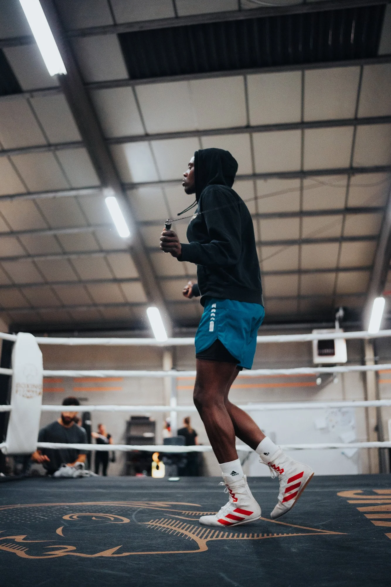 A man in boxer gear, including shorts and boxing shoes, is jumping rope in a gym boxing ring.