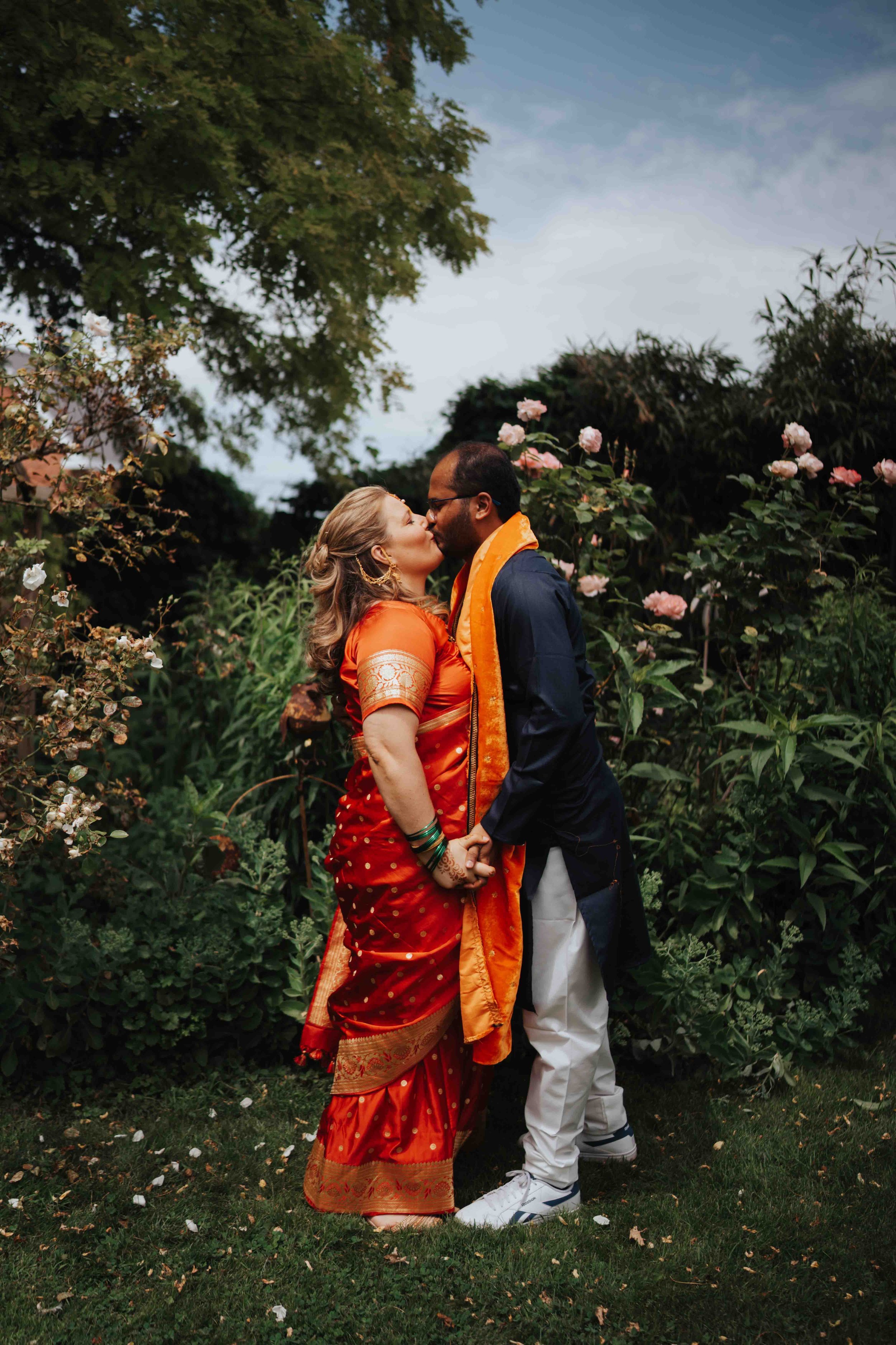 A couple dressed in traditional Indian attire sharing a kiss in a garden surrounded by blooming roses.