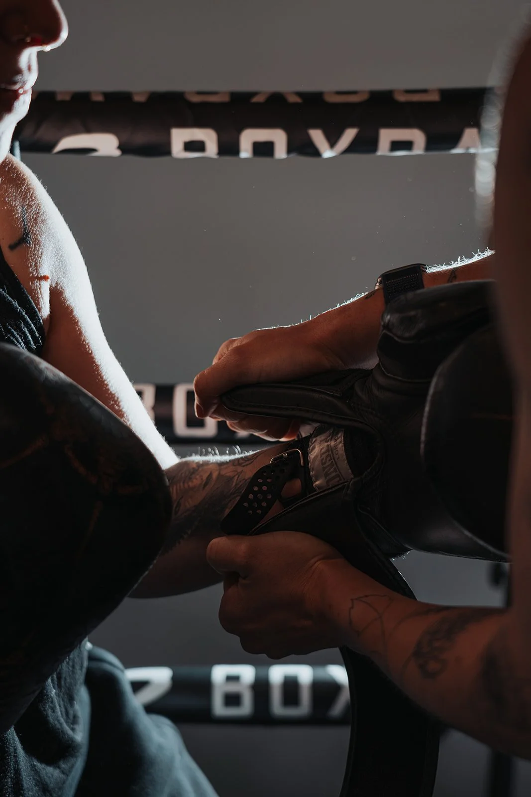 A person with tattoos is putting on and fastening her boxing gloves, with another person helping. There is a banner with the word 'BOX' visible in the background, suggesting a boxing gym or training environment.