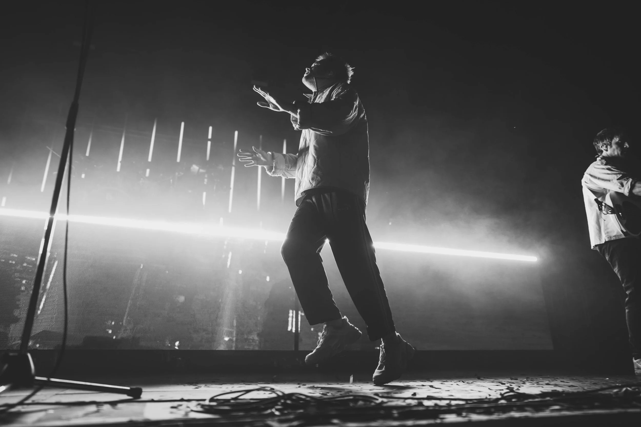 Rou Reynolds of Enter Shikari dancing on stage with neon lights in the background, in black and white.