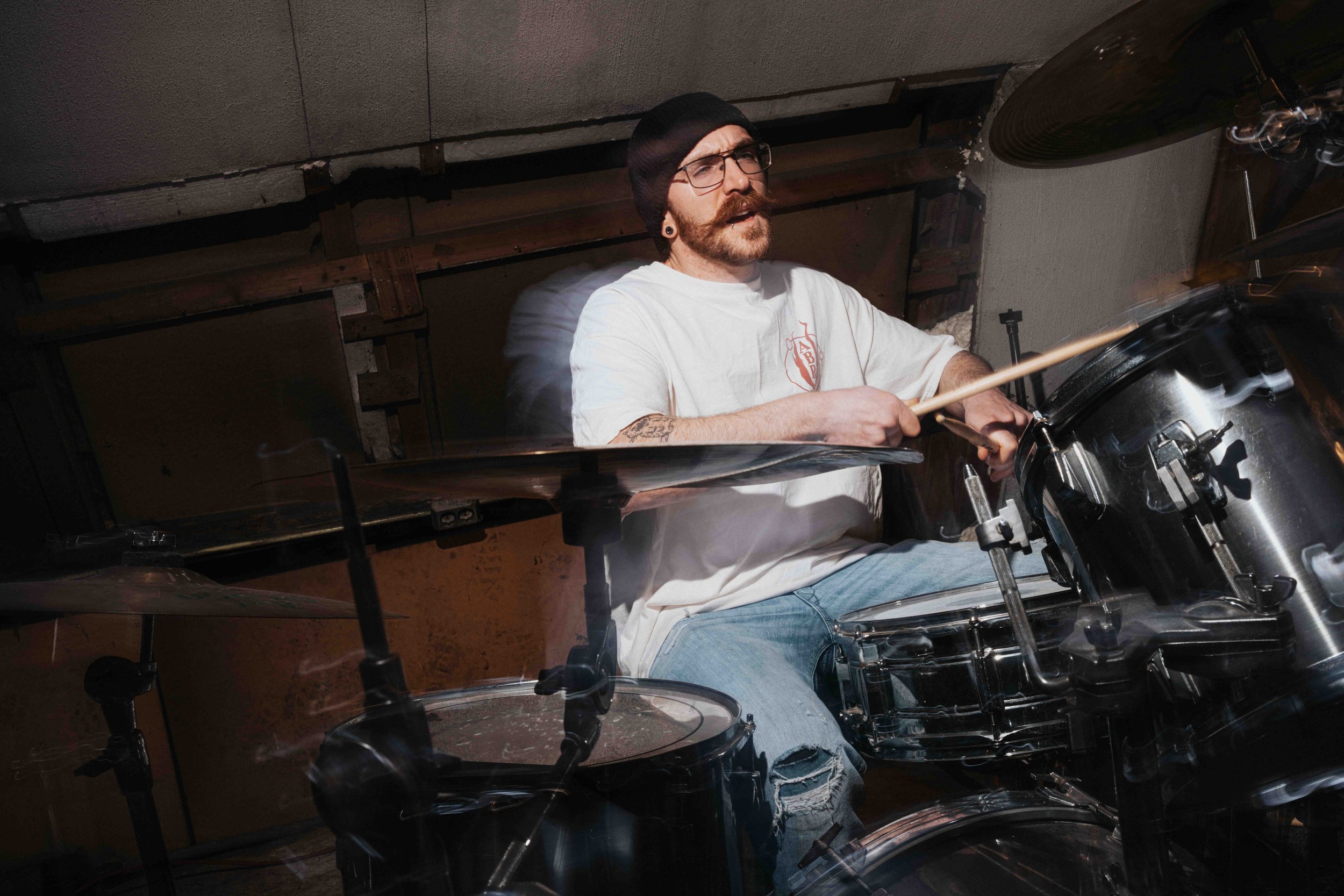 A man with glasses and a beard playing the drums in a garage or basement. He is wearing a white t-shirt, a black beanie, and jeans, with tattoos on his arm.