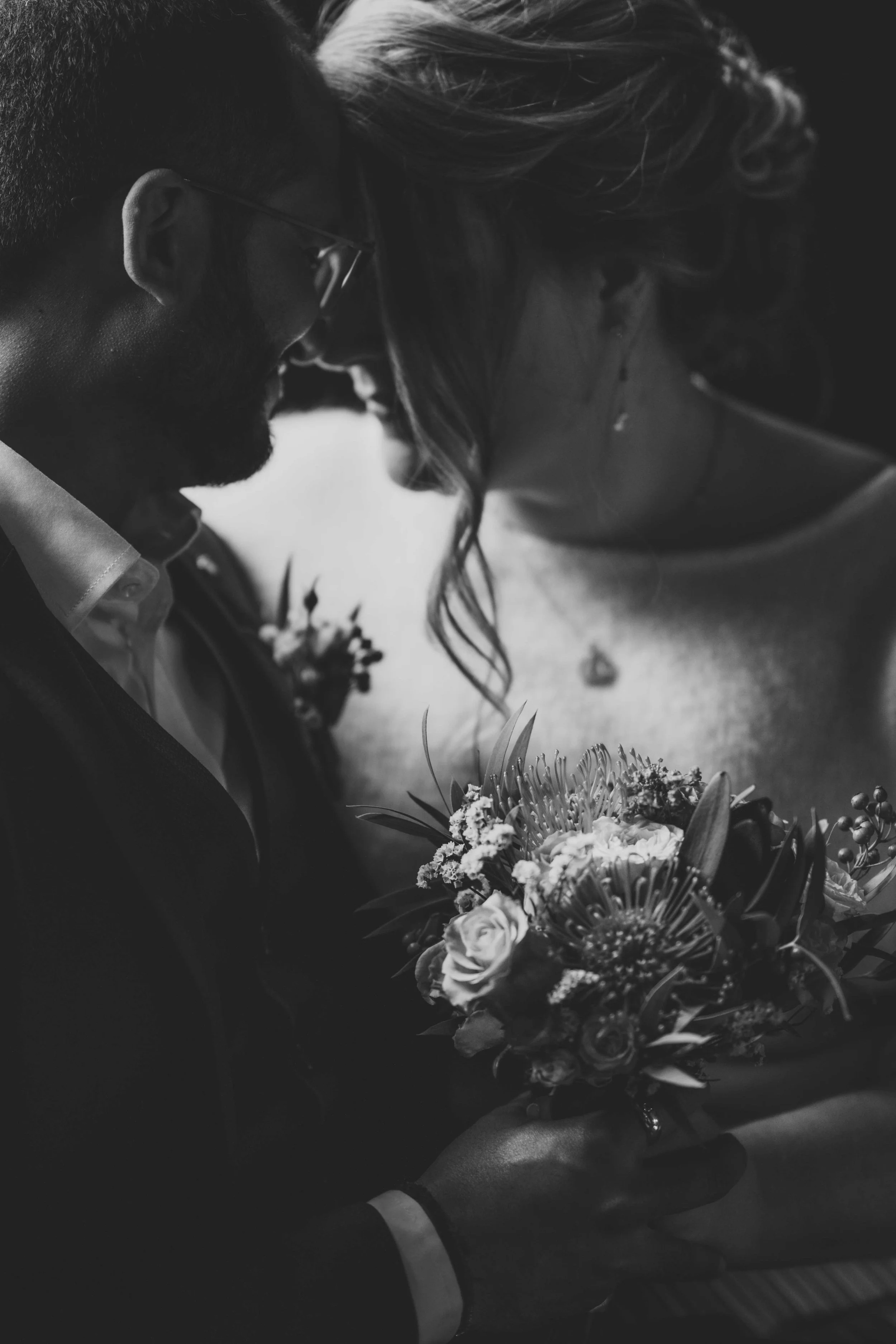 A black and white photo of a couple, likely newlyweds, holding a bouquet of flowers together. The man has a beard, glasses, and is wearing a suit jacket. The woman has wavy hair, a necklace, and a dress. They are touching foreheads and smiling softly