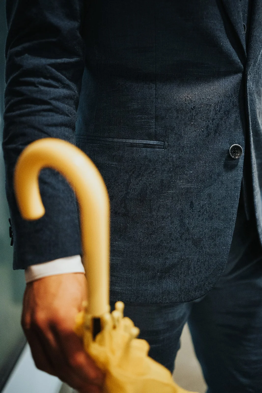 Close-up of a person in a dark suit holding a yellow umbrella, with focus on the suit and umbrella handle.