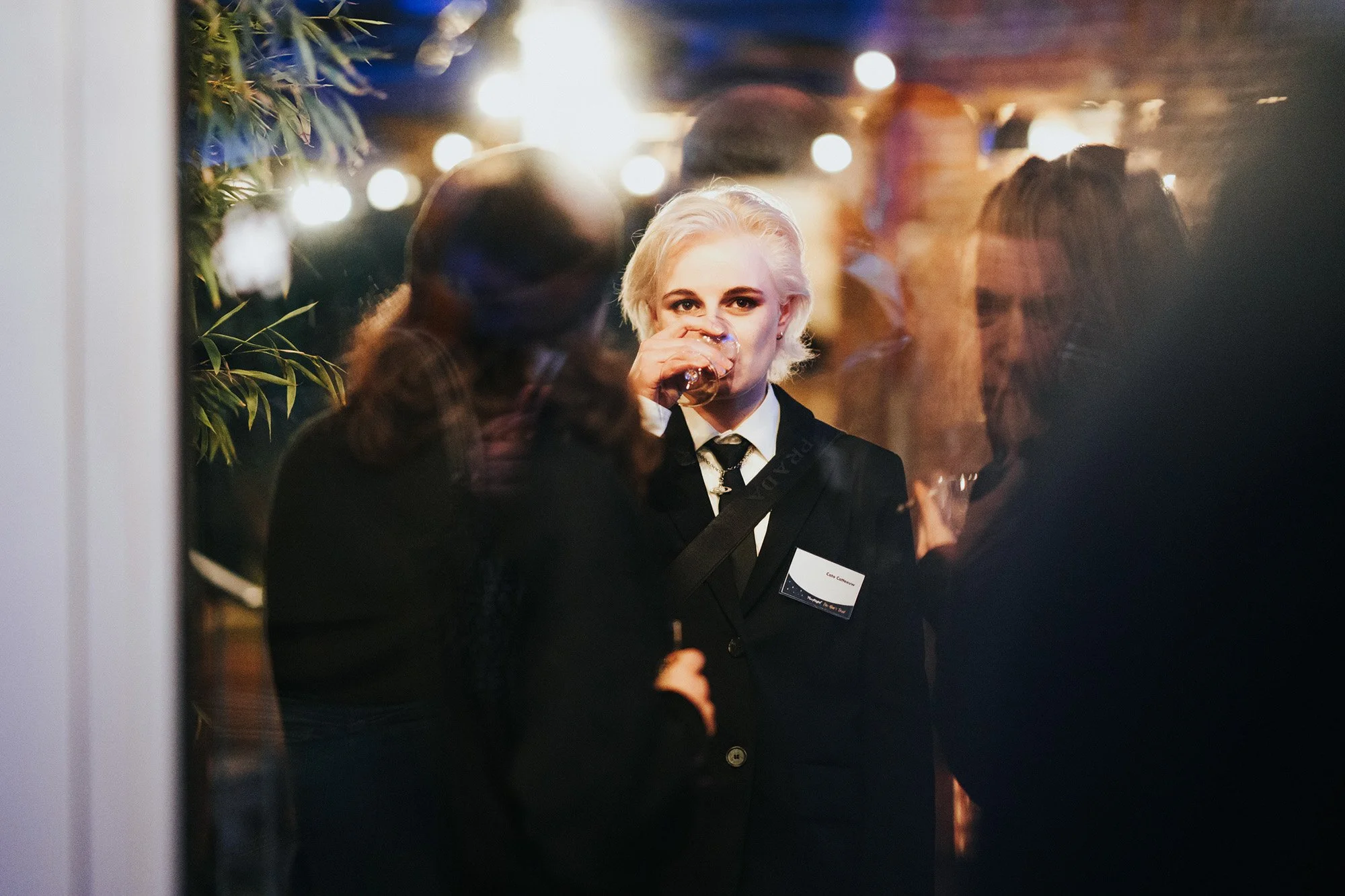 A woman with platinum blonde hair wearing a black suit and white shirt, drinking from a glass, at an indoor social gathering.