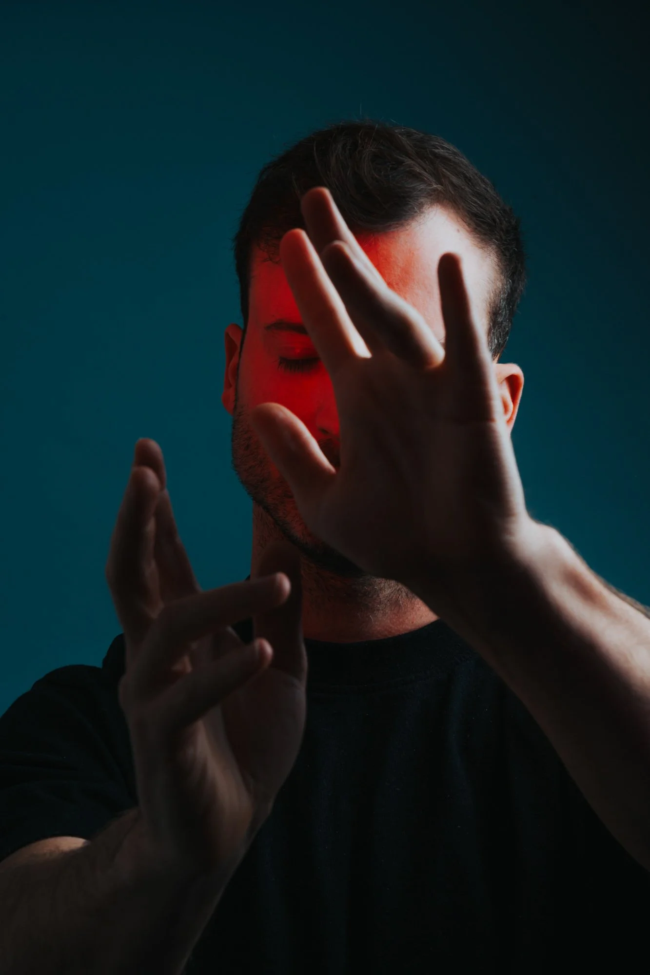 A man with short dark hair and a beard (Pianist Pieter Savenberg), wearing a black shirt, is covering his face with his right hand and partially with his left hand. The background is dark with a red light shining on his face.
