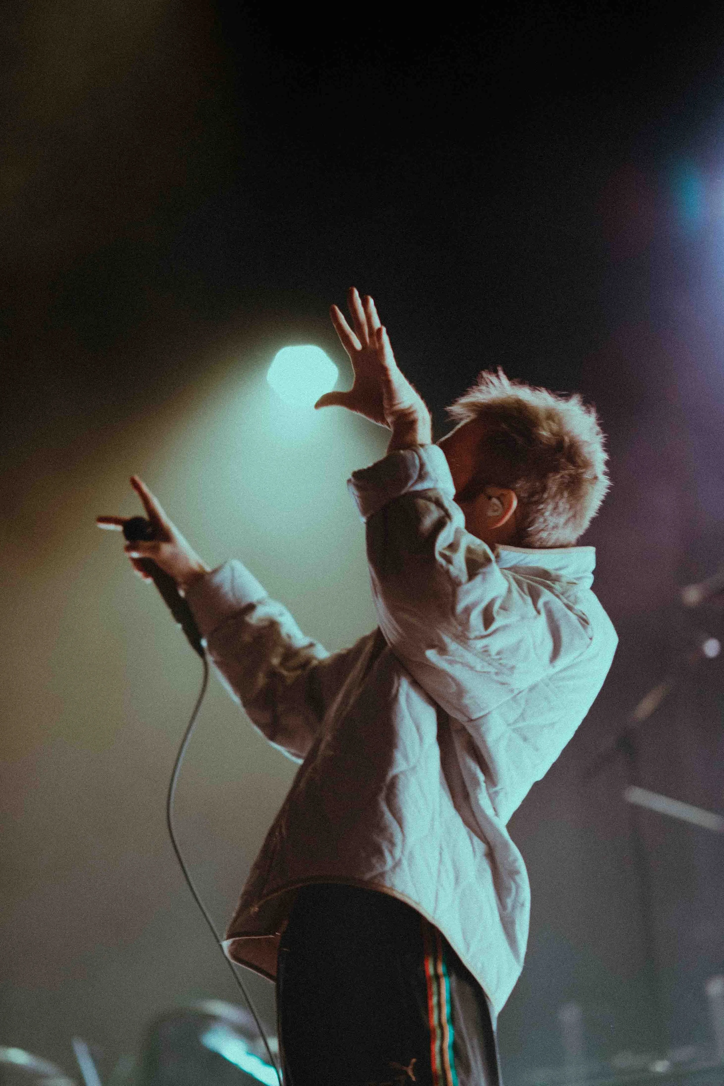 Performer Rou Reynolds of Enter Shikari singing on stage, holding a microphone, wearing a light-colored jacket, with stage lights creating a backlit effect.