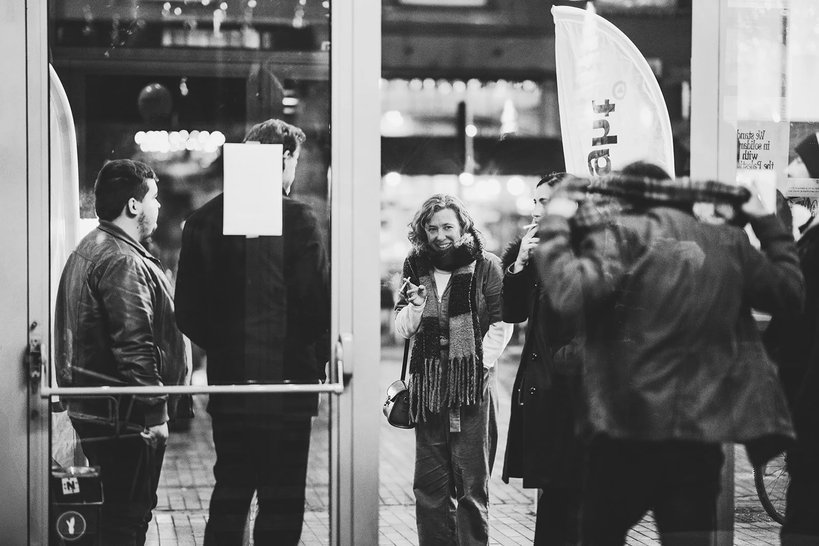 A group of people standing outside a glass door on a city street at night. One woman in the center, smiling and holding a cigarette, faces the camera with others engaged in conversation around her.