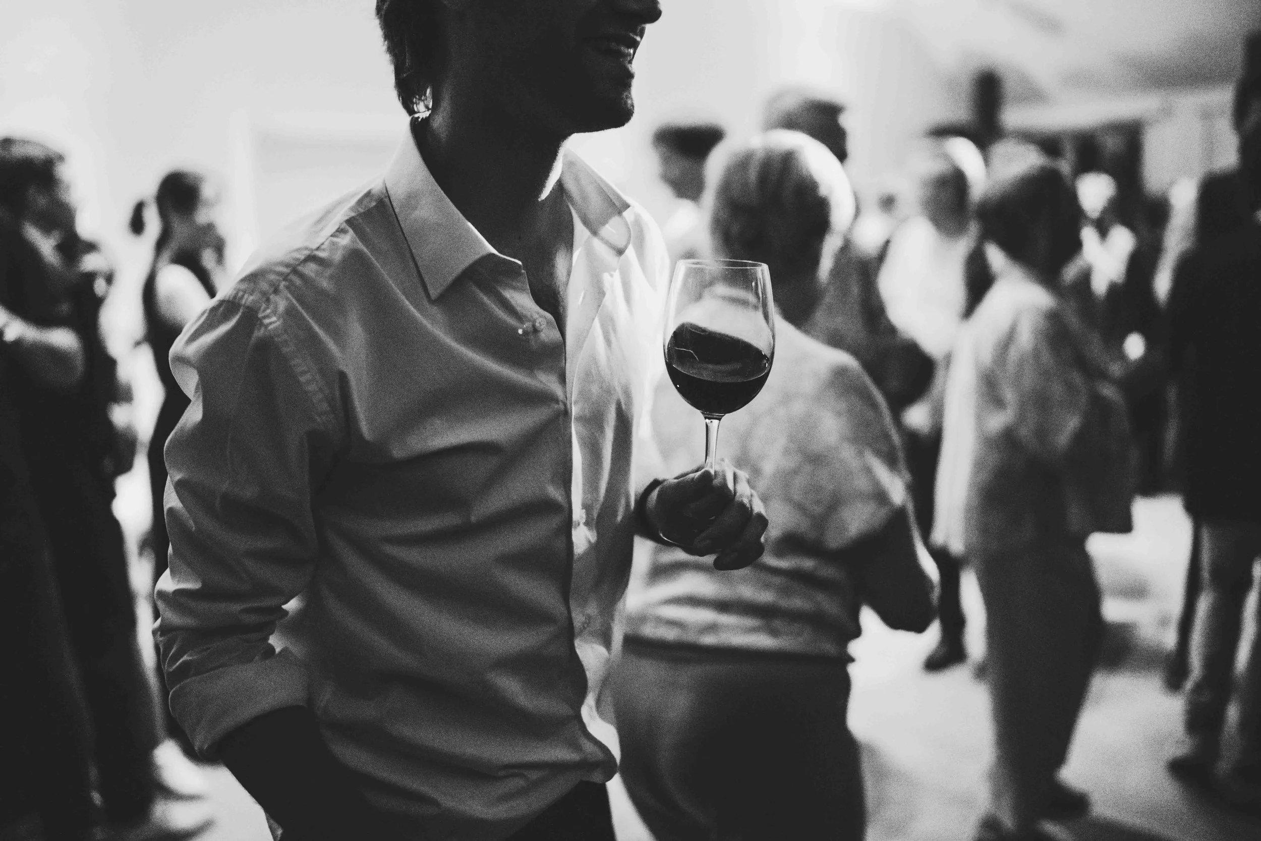 Man holding a glass of red wine at a social gathering, with people mingling in the background.