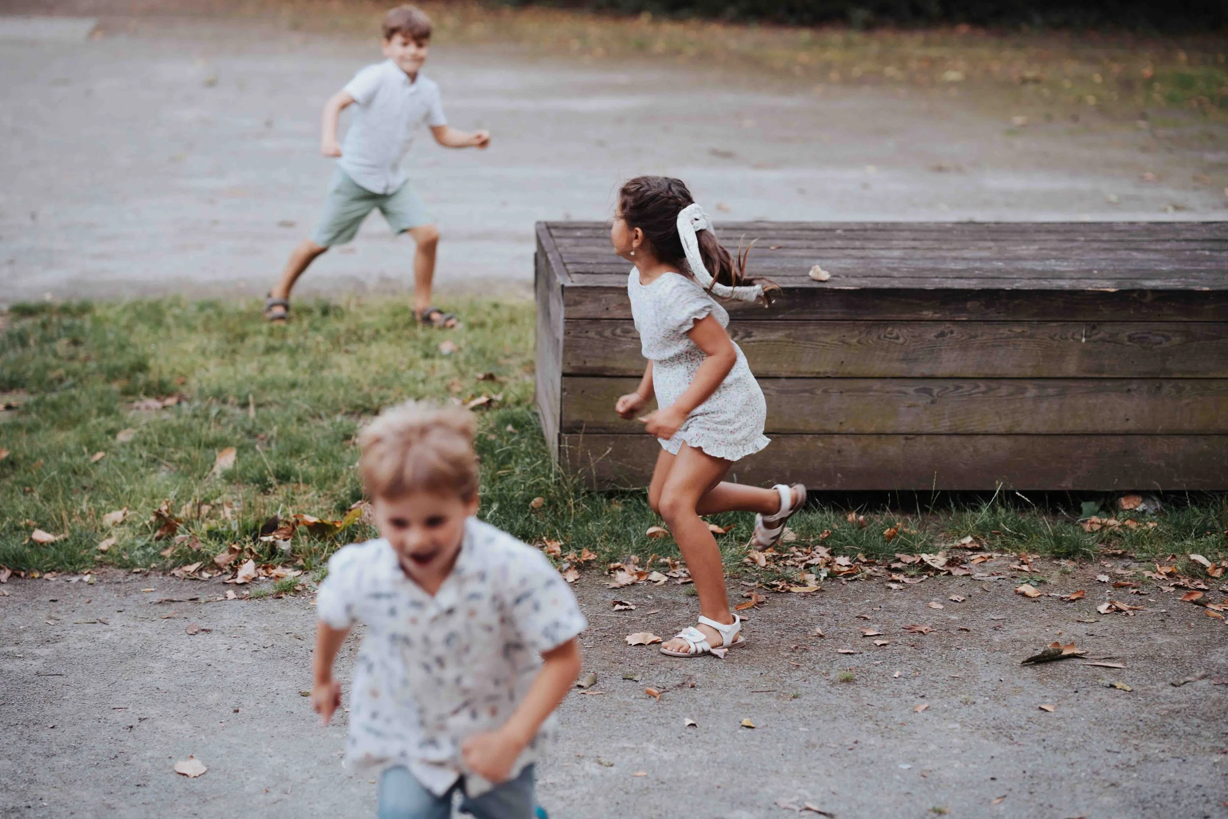 Children playing and running outdoors near a wooden structure and fallen leaves on the ground.