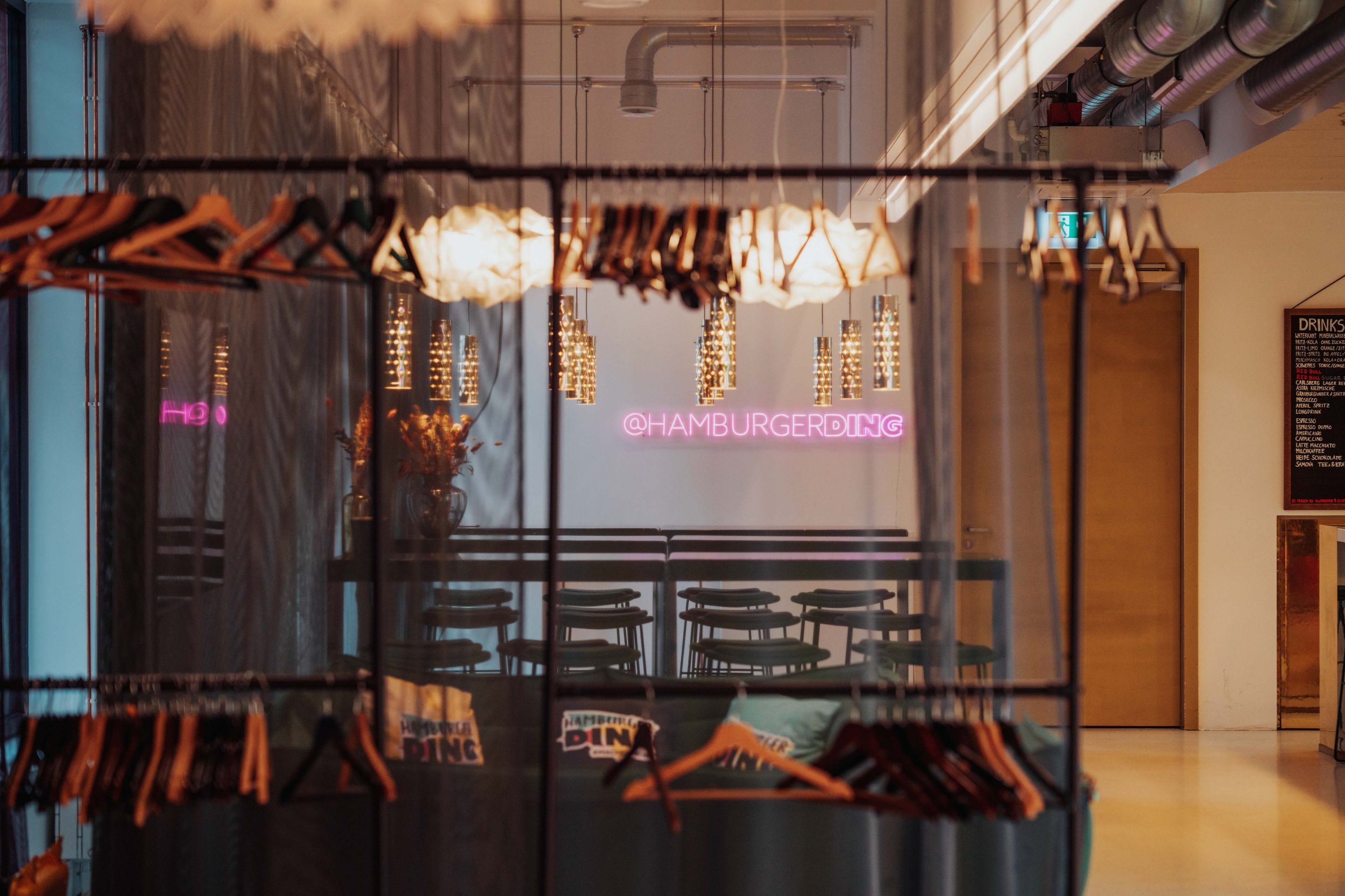 Interior of a restaurant or cafe with a neon sign that reads '@HAMBURGERDING'. The space features hanging pendant lights, a row of stools, and a promotional rack with hangers in the foreground.
